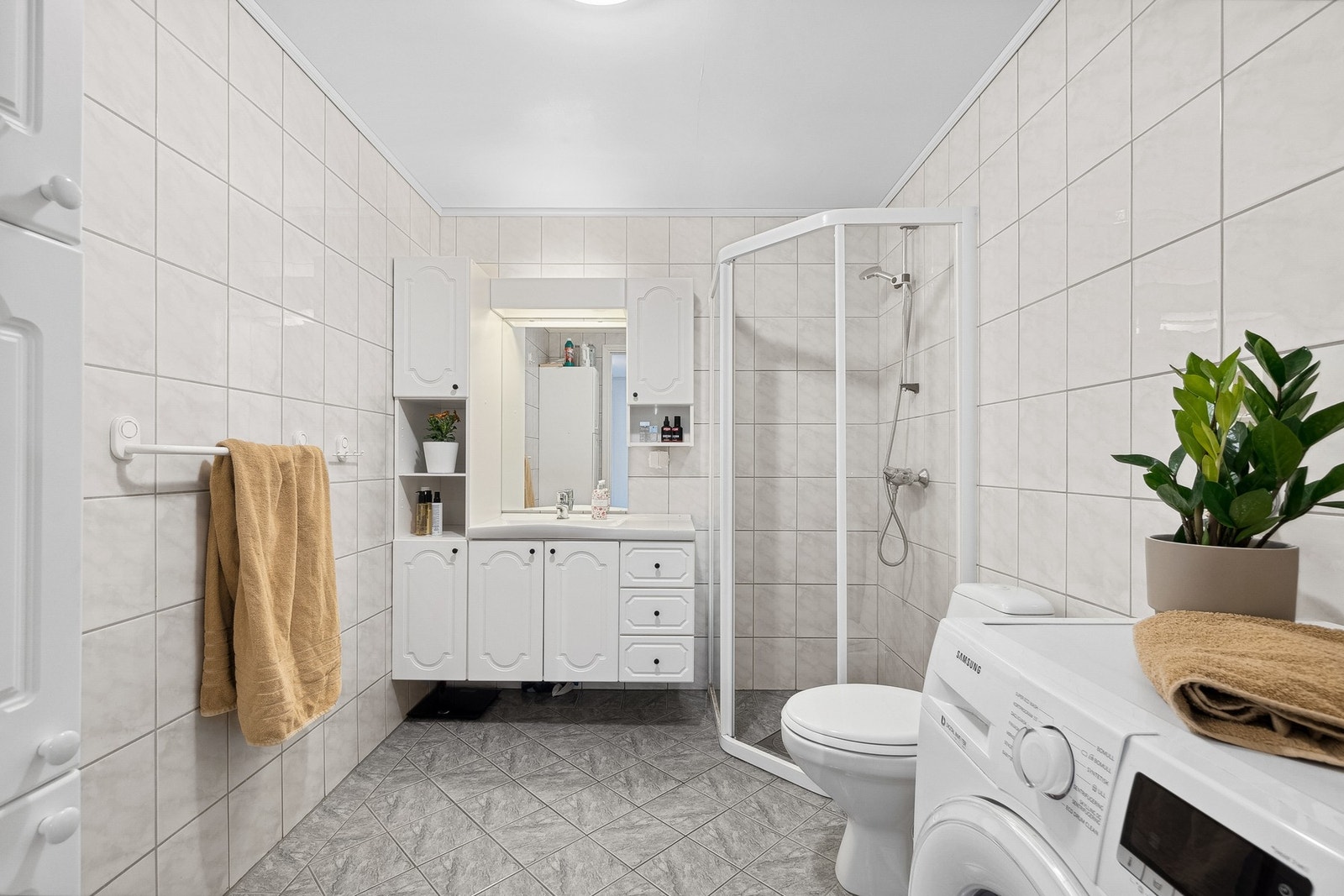 Romslig flislagt bad med lys innredning, dusjhjørne, Wc og opplegg for vaskemaskin. Galleribilde