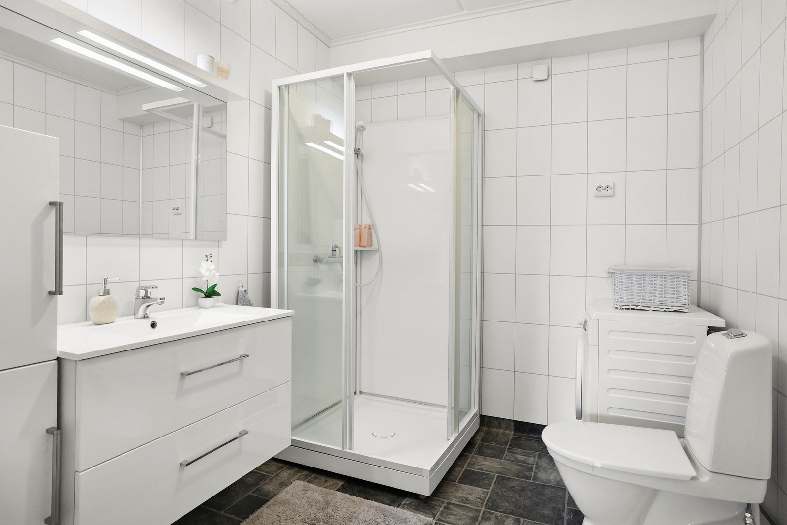 Badet inneholder vegghengt servantinnredning og speil med overlys, dusjkabinett, gulvstående toalett og opplegg for vaskemaskin. Galleribilde