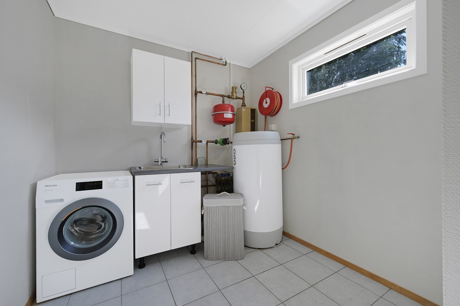 Eget vaskerom med fliser på gulv og malte vegger. Rommet har opplegg for vaskemaskin, tørketrommel og det er en utslagsvask. Galleribilde