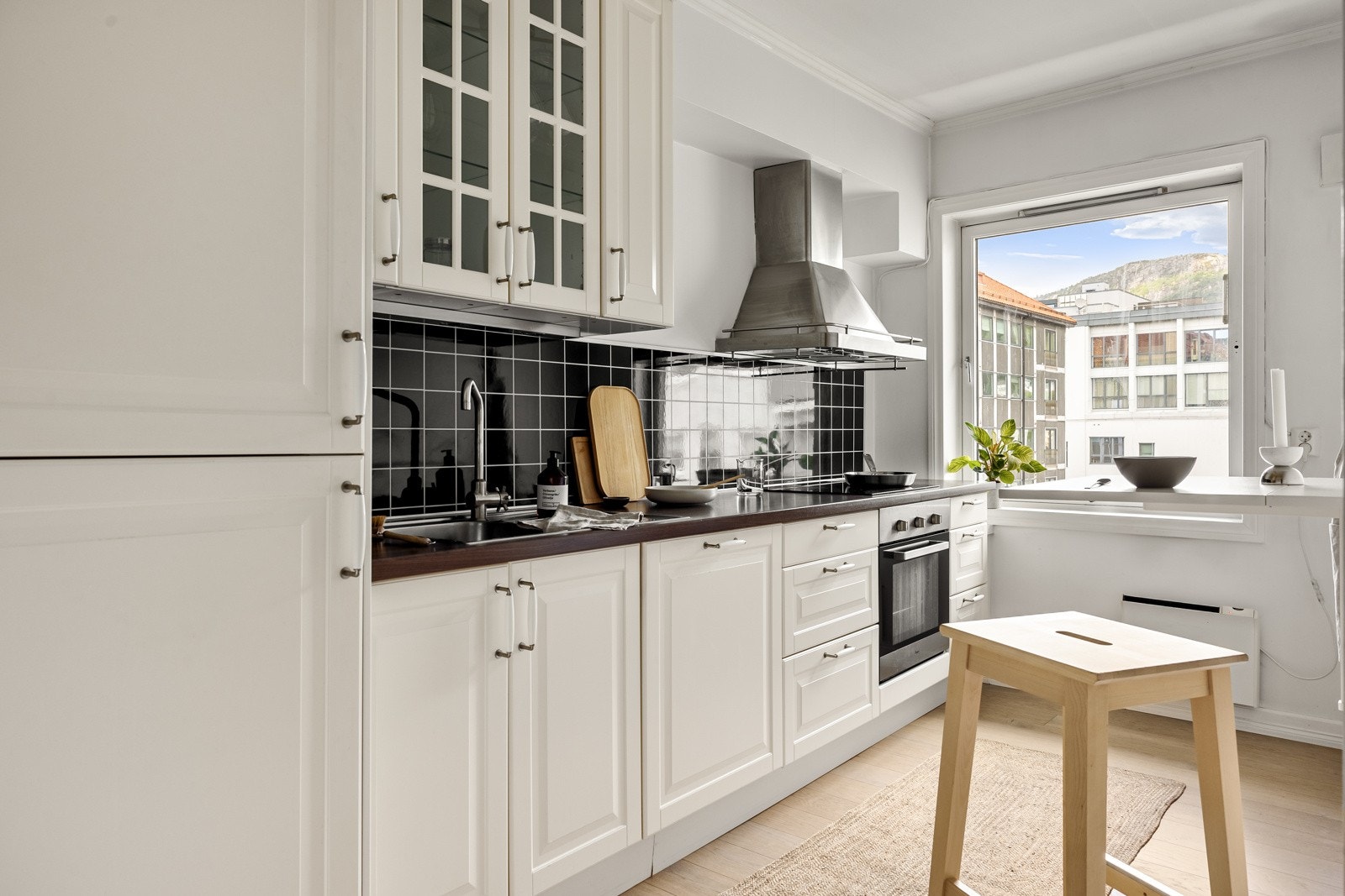 Romslig kjøkken med profilerte hvite fronter, glasskap og laminat benkeplate med nedfelt vask av stål og veggmontert ventilator. Galleribilde