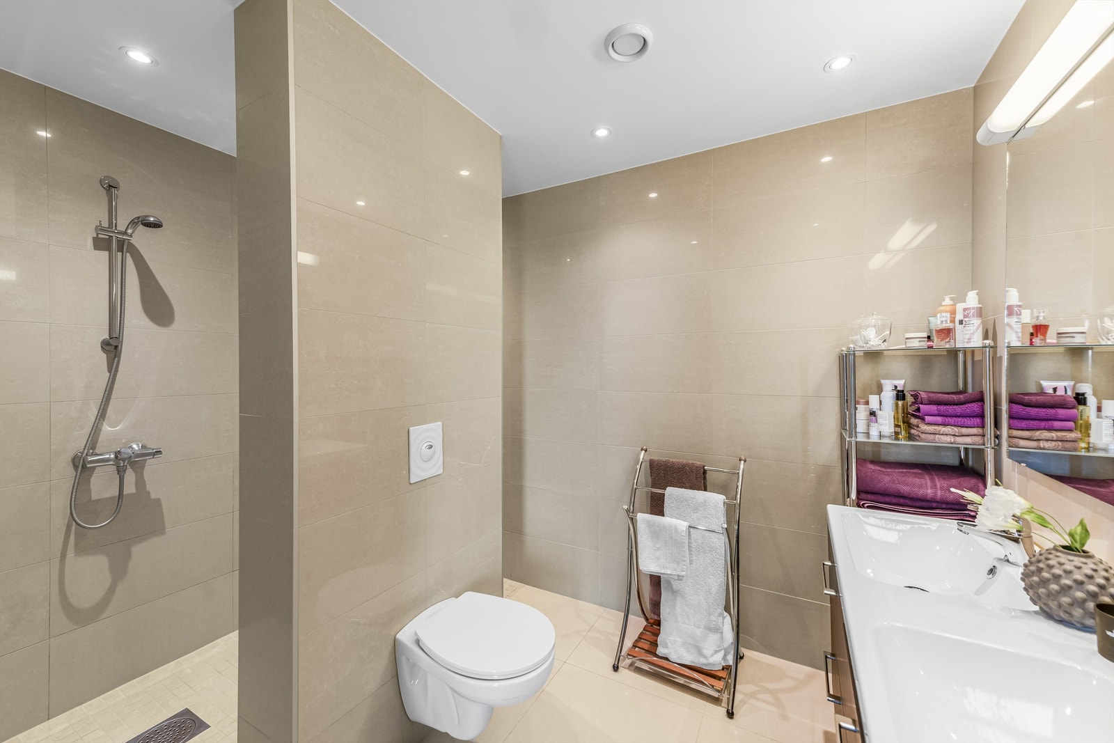 Beige fliser på gulv og vegger, gulvvarme og downlights. Innredningen består av en heldekkende dobbelservant på mørk skuffeseksjon, et heldekkende speil med overlys, et veggmontert toalett, og to dusjnisjer bak flislagt skillevegg. Galleribilde