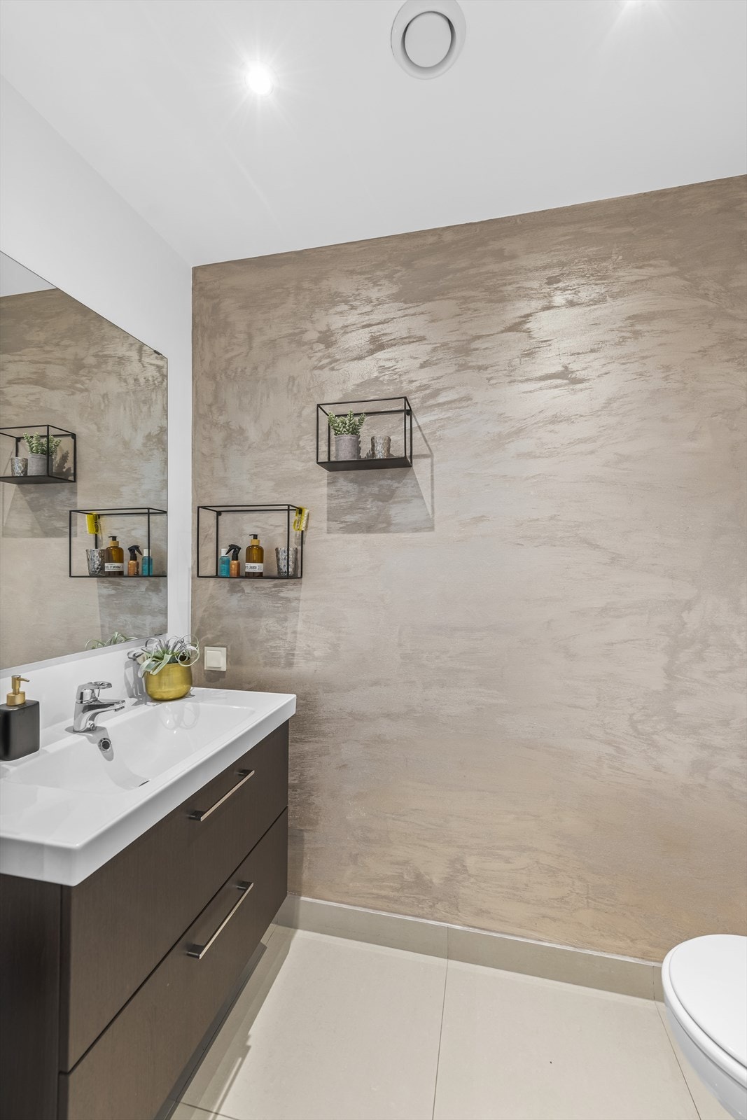 Toalettrommet i samme etasje har lyse fliser på gulv med gulvvarme, lysmalte veggflater, fondvegg med kalkmaling og listefritt tak med downlights. Innredningen består av en heldekkende servant på mørk skuffeseksjon, speil og veggmontert toalett. Galleribilde