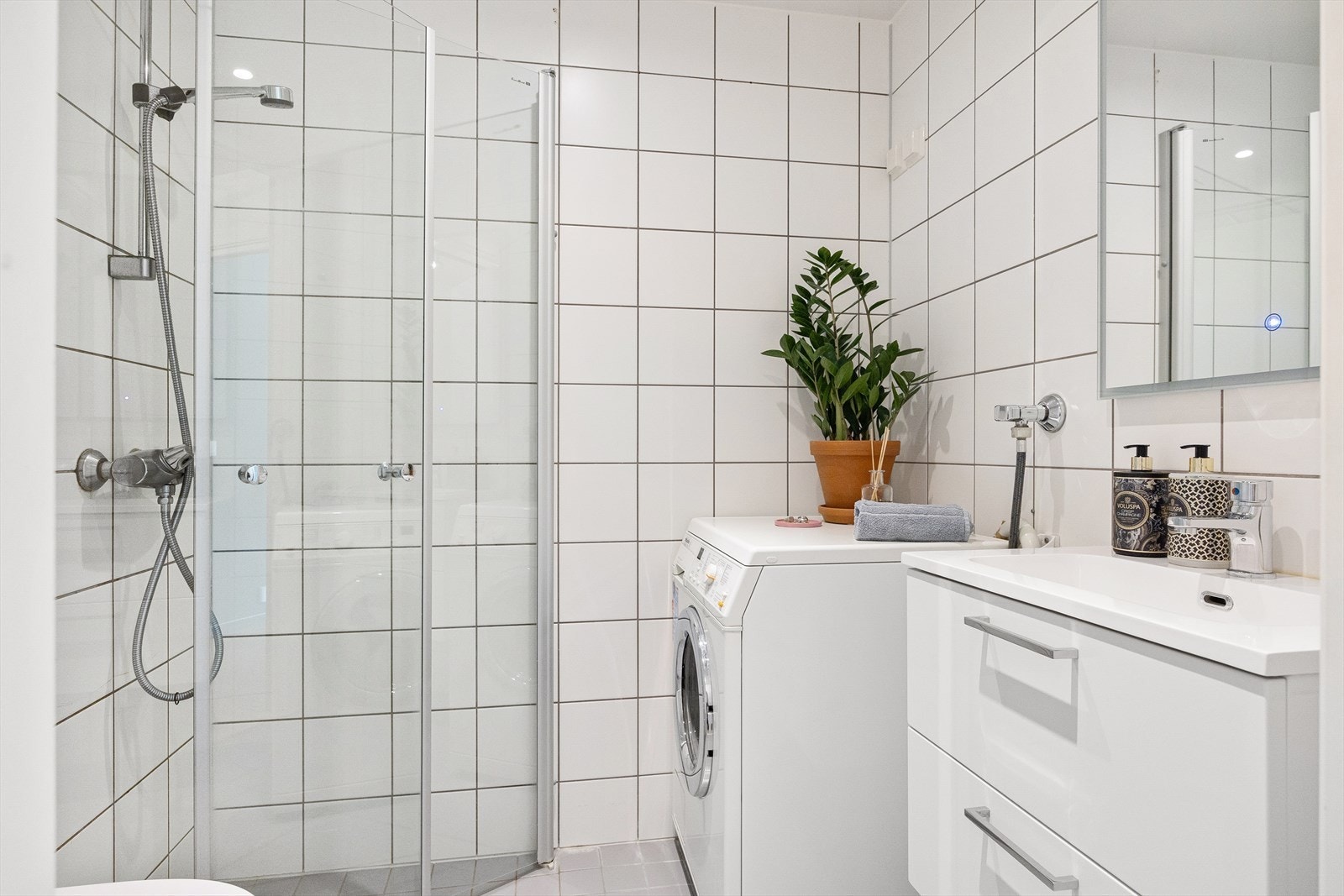 Pent baderom med flis på gulv og vegg. Det er etablert servantskap med nedfelt servant, speil med innebygd berøringslys, klosett med innebygd sisterne og dusjhjørne med innfellbare dusjdører i herdet glass. Galleribilde