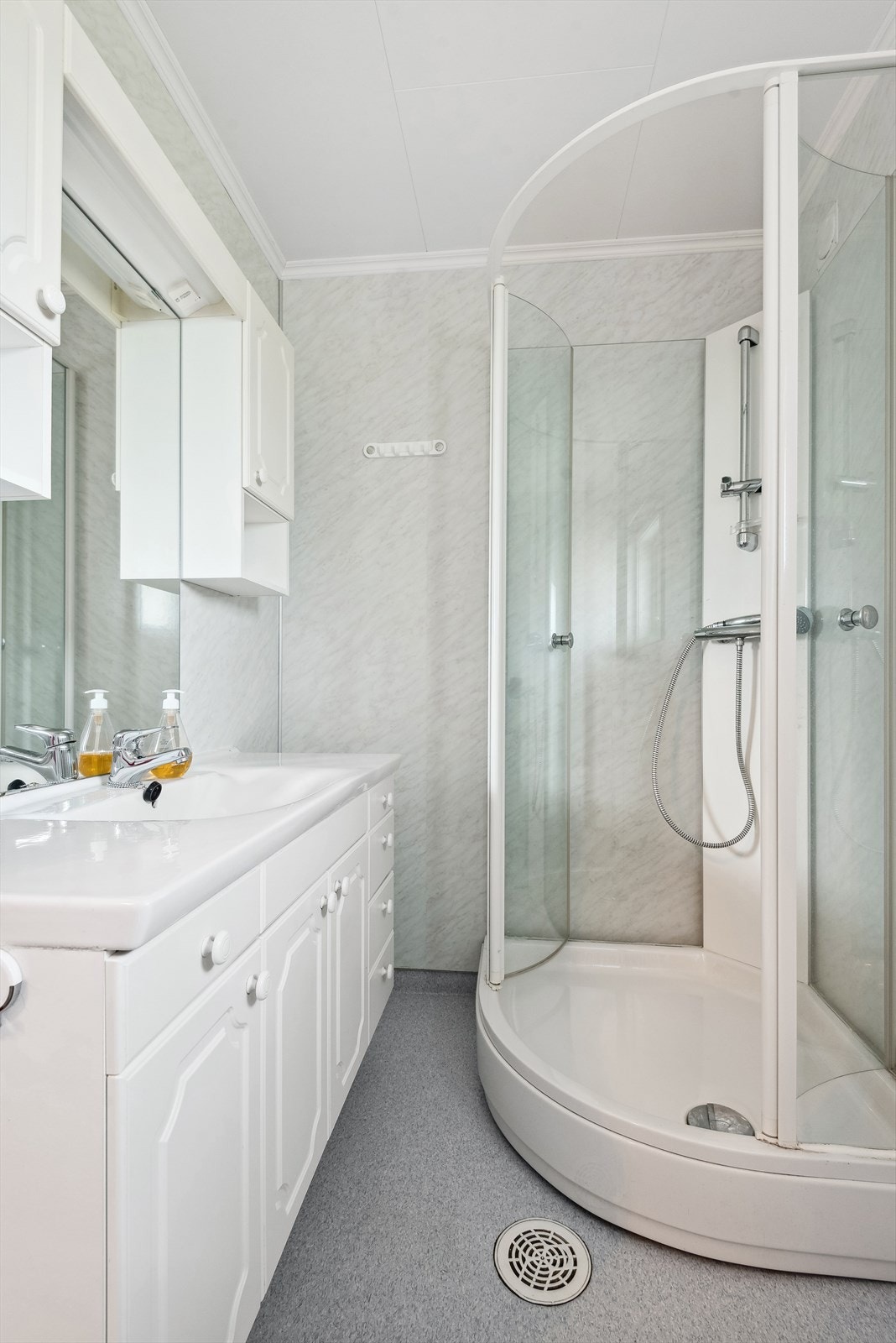 Badet er utstyrt med dusjkabinett, gulvmontert toalett og en romslig baderomsinnredning med underskap, skuffer og heldekkende servant. På veggen i overkant er det veggskap, speil, lys og elektrisk uttak. Galleribilde