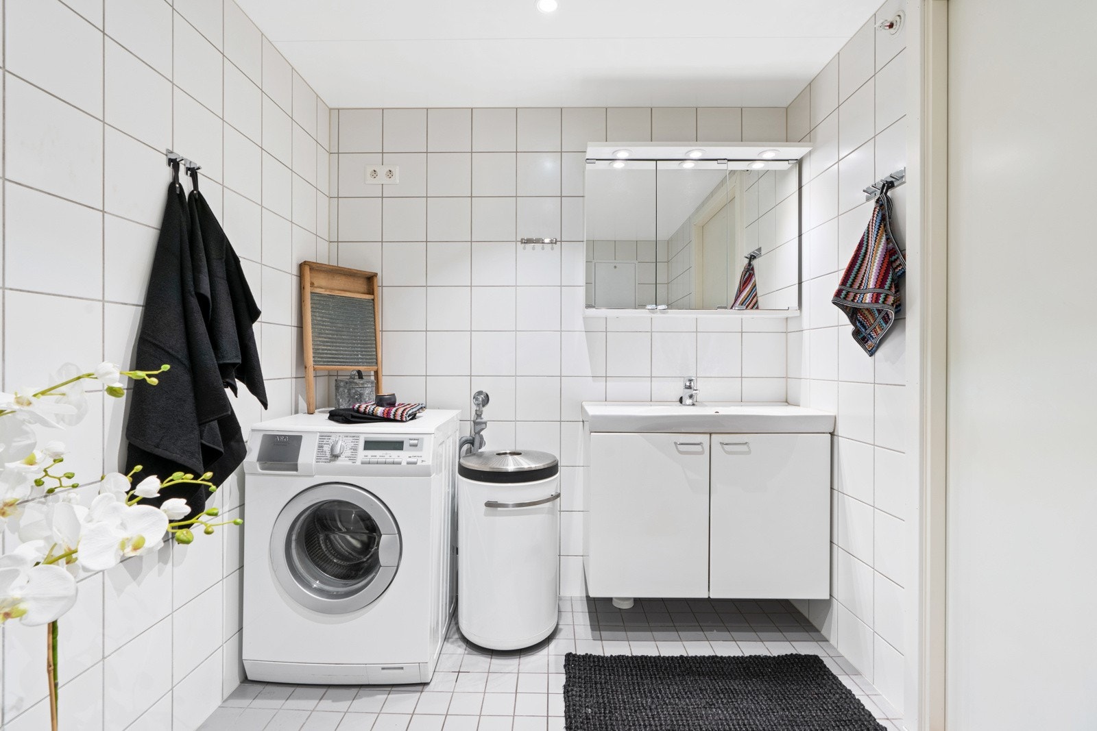 Begge har grå fliser på gulv med gulvvarme, hvite fliser på veggene og downlights i himling. Dette badet har opplegg for vaskemaskin. Galleribilde
