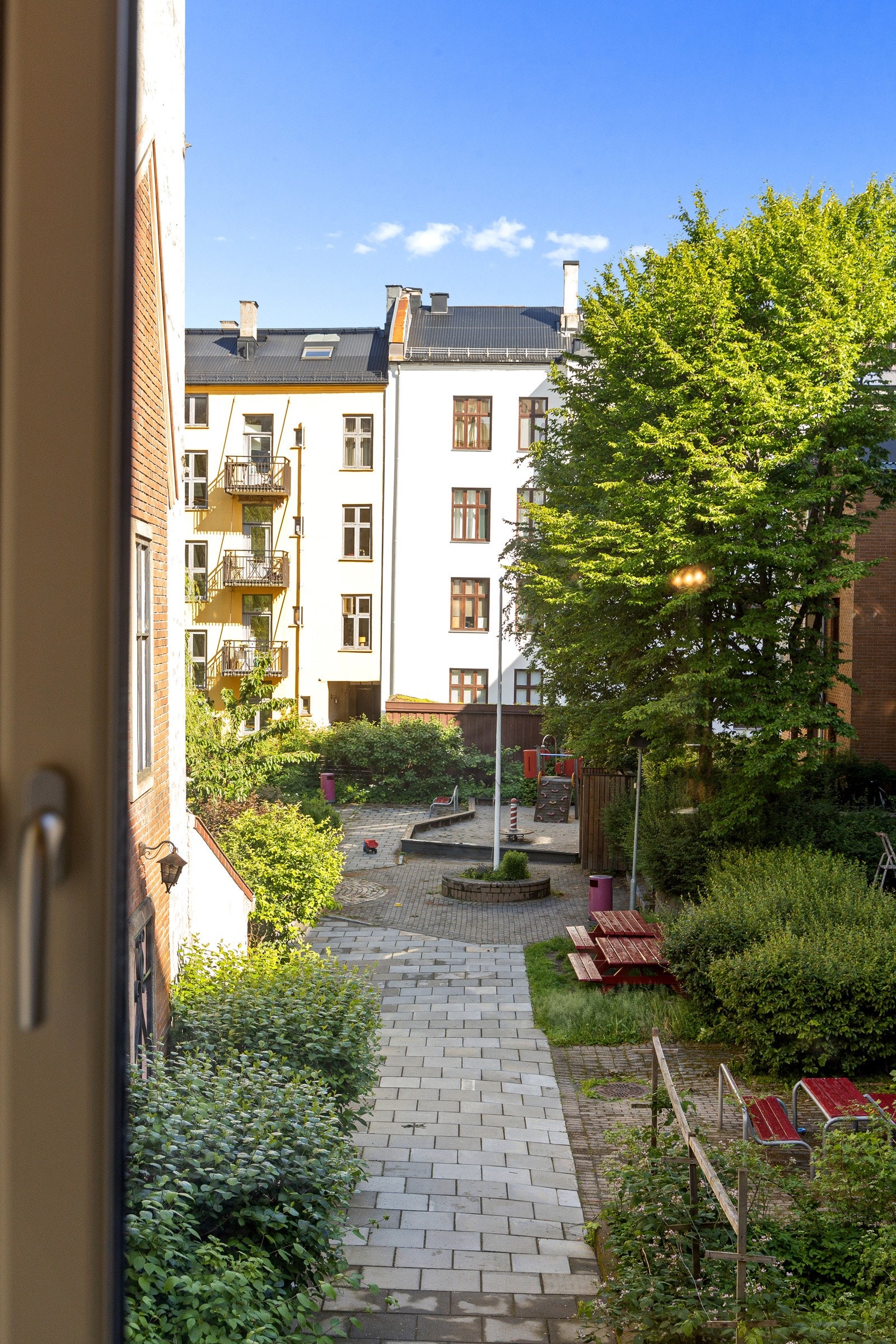 Utsikt fra kjøkkenvinduet mot stor bakgård med lekeplass, grill og sittegrupper. Galleribilde