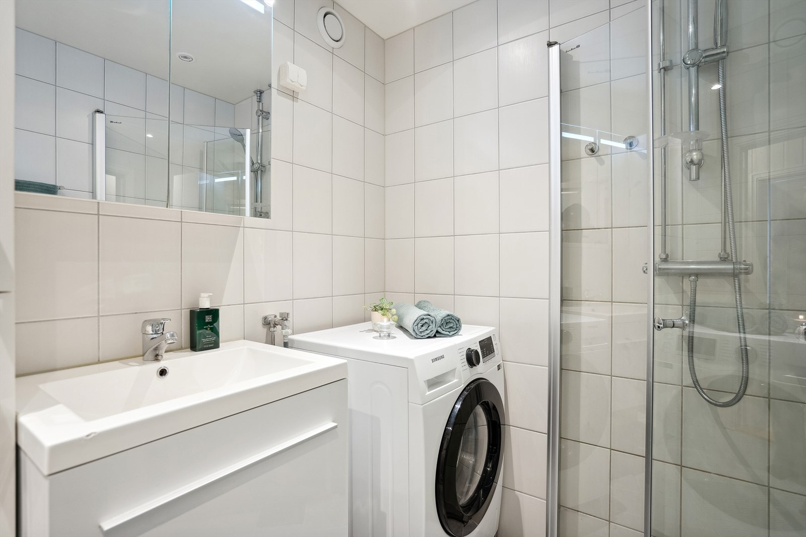 Speil med lys, høyskap og dusj i hjørnet bak svingbare glassvegger samt opplegg for vaskemaskin Galleribilde