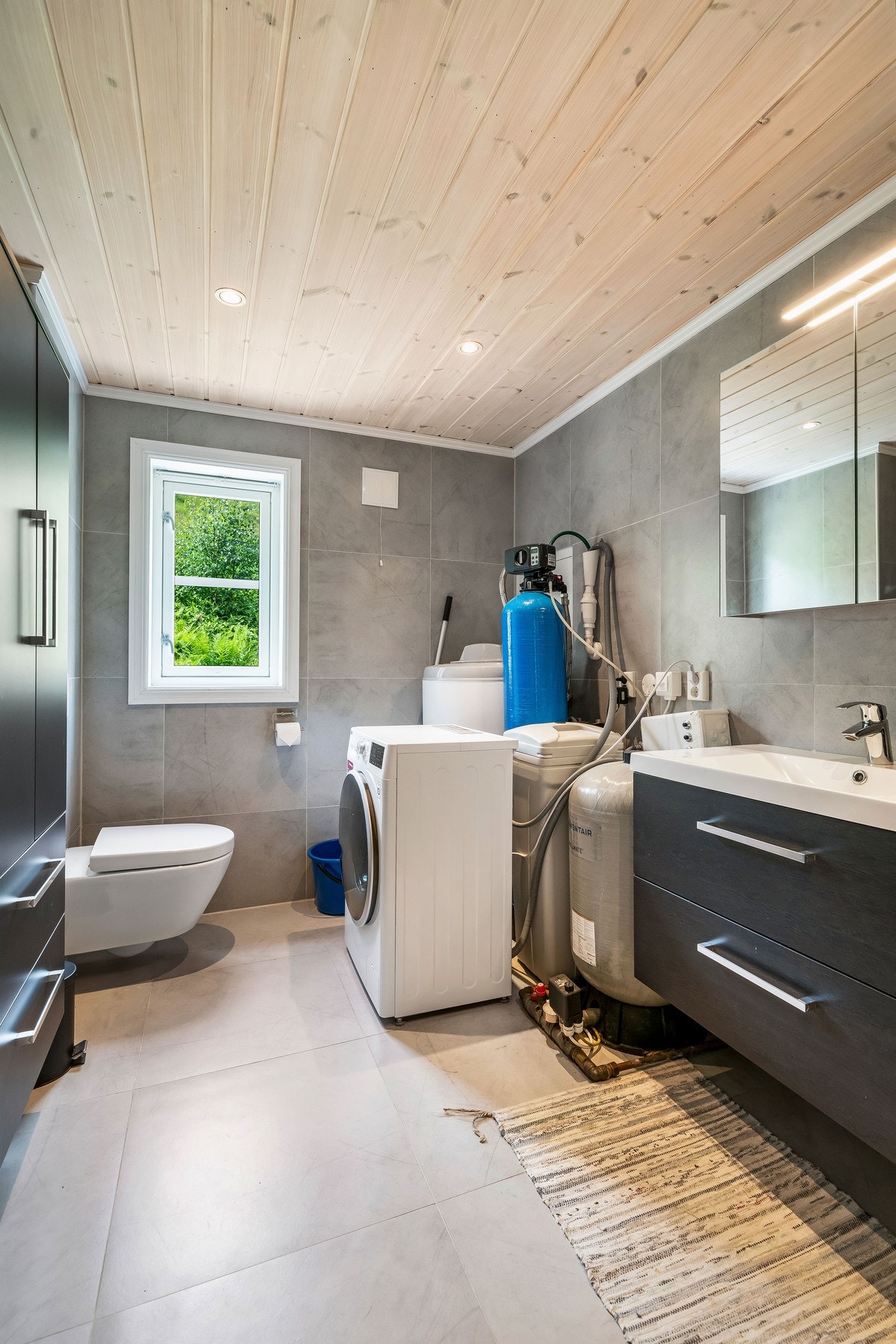 Baderommet er utstyrt med dusjhjørne med innfellbare glassvegger, to-greps blandebatteri, takdusj og hånddusj. Vegghengt servantskap med heldekkende servant med ett-greps blandebatteri. Overskap med speil, høyskap, samt vegghengt toalett. Opplegg for vask Galleribilde