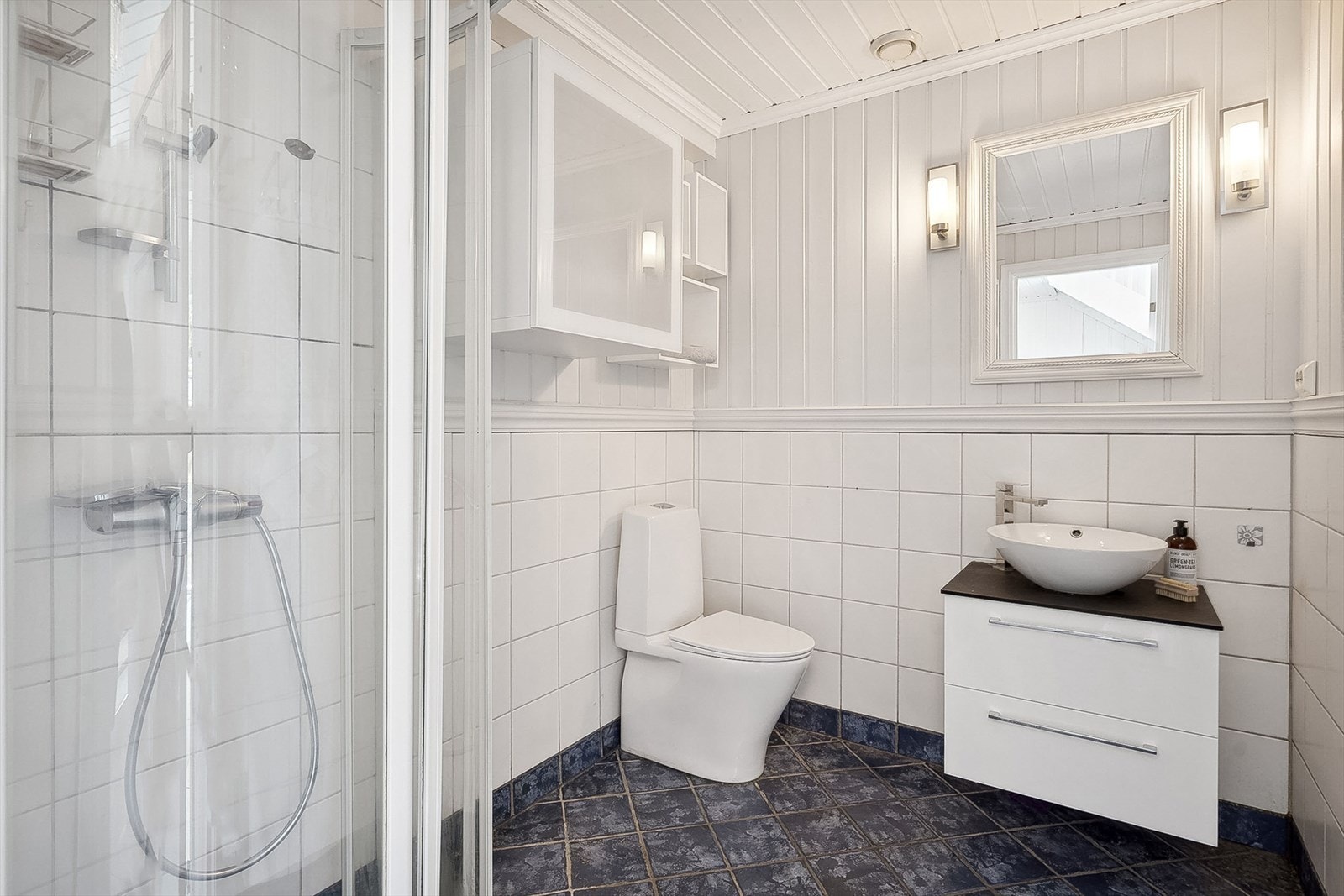 Boligen inneholder hele tre bad - ett i hver etasje. Her fra badet beliggende i 1.etasje med innredning fra 2019 med bolleservant på hvit skuffeseksjon, speil med belysning, et skap, et toalett og et eldre dusjhjørne. Galleribilde