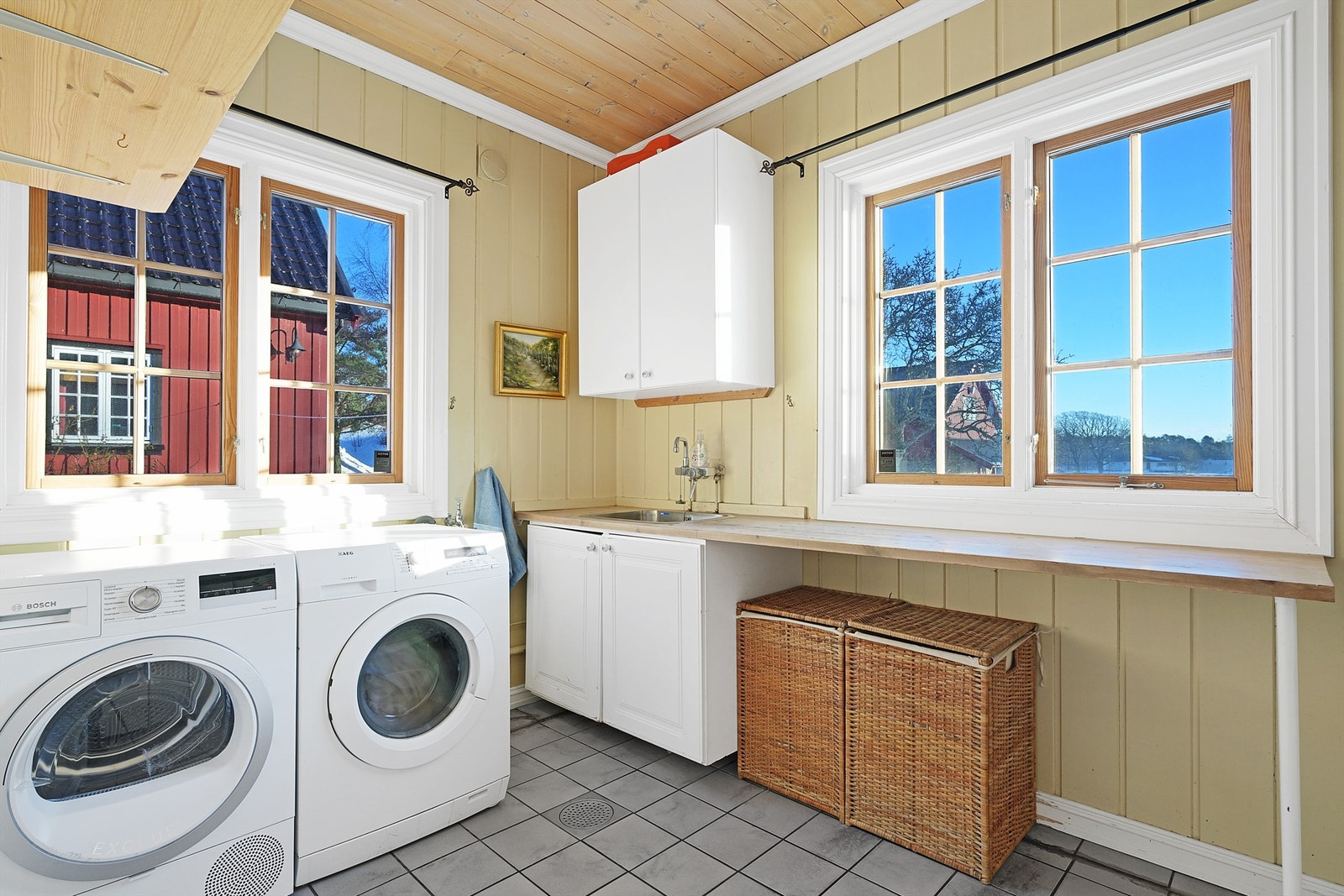 Praktisk vaskerom med varmekabler malt trepanel på vegger, trepanel i tak og opplegg for vaskemaskin og tørketrommel. Her er det enkel innredning med utslagsvask nedfelt i benkeplate. Galleribilde