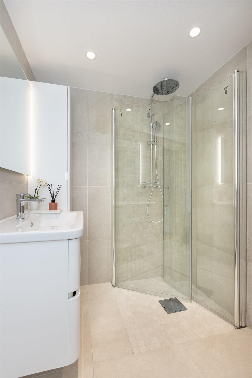 Vegghengt servantskap, ovenpåliggende servant, speil med belysning på vegg over servant, vegghengt høyskap ved siden av servant, dusjsone med innfellbare dører av glass, dusjarmatur tilkoblet hånddusj og regndusj, samt vegghengt toalett Galleribilde