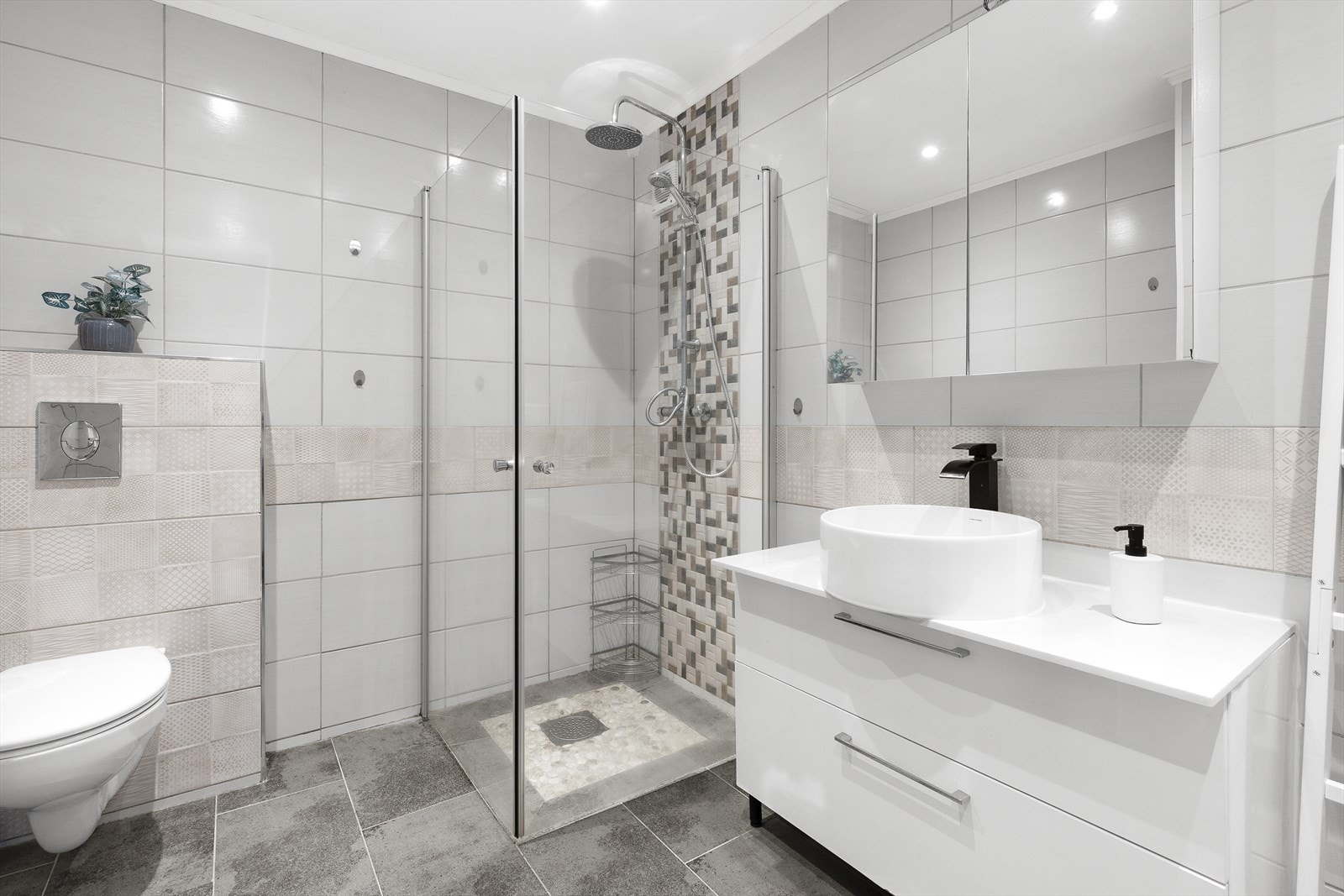 Flislagt og innholdsrikt baderom innredet med servant på skuffeinnredning, overskap med speildører, dusjhjørne, vegghengt wc og varmtvannsbereder. Galleribilde