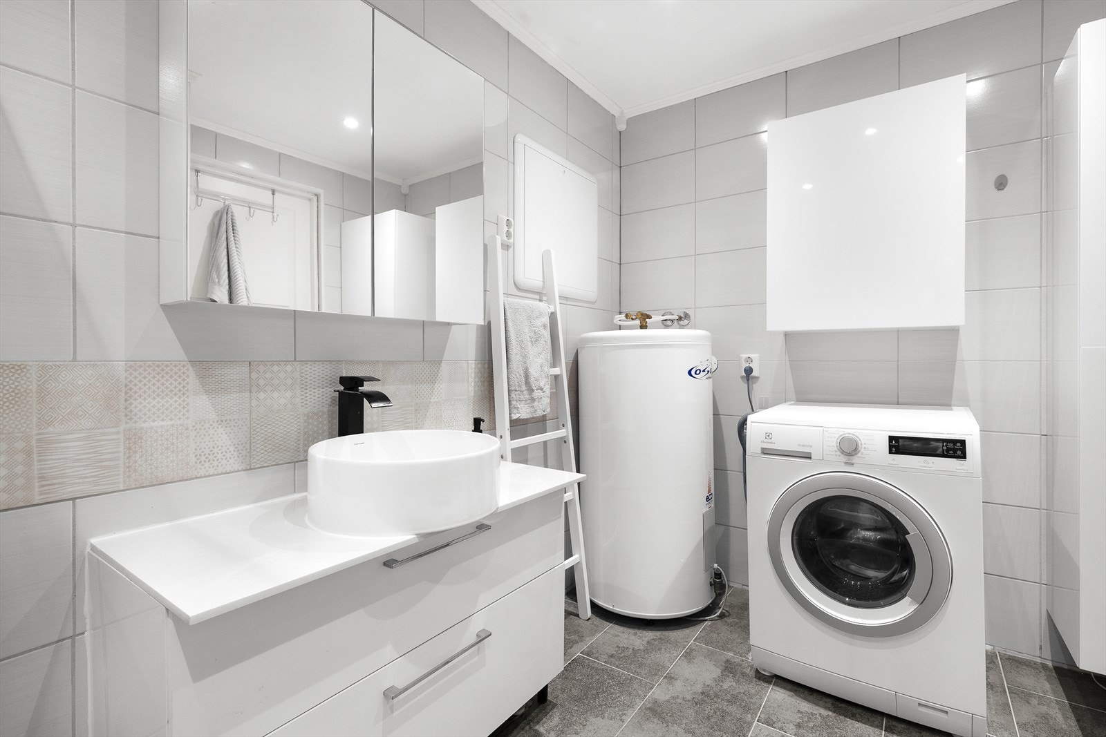 Flislagt og innholdsrikt baderom innredet med servant på skuffeinnredning, overskap med speildører, dusjhjørne, vegghengt wc og varmtvannsbereder. Galleribilde