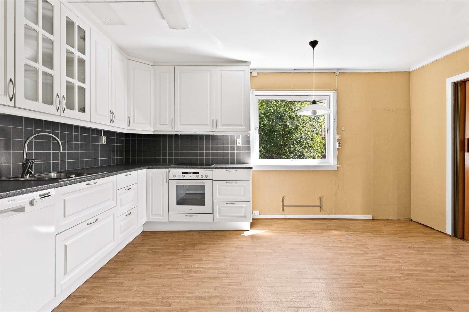 Kjøkkeninnredning med profilerte hvite fronter, laminert benkeplate med nedfelt dobbel stålkum med ettgreps blandebatteri og overskap med glass. Det er montert kitchenboard på veggen mellom over- og underskapene. Galleribilde