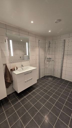 Stort lyst bad med fliser på gulv og baderomsplater på vegg, med opplegg til vaskemaskin og varmtvannsbereder. Galleribilde