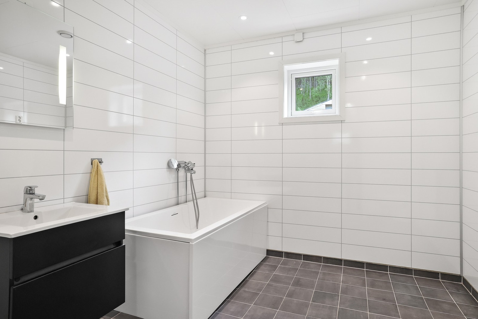 Det er en mørk innredning med nedfelt servant, veggmontert toalett, badekar og opplegg for vaskemaskin. I tillegg er det balansert ventilasjon. Galleribilde