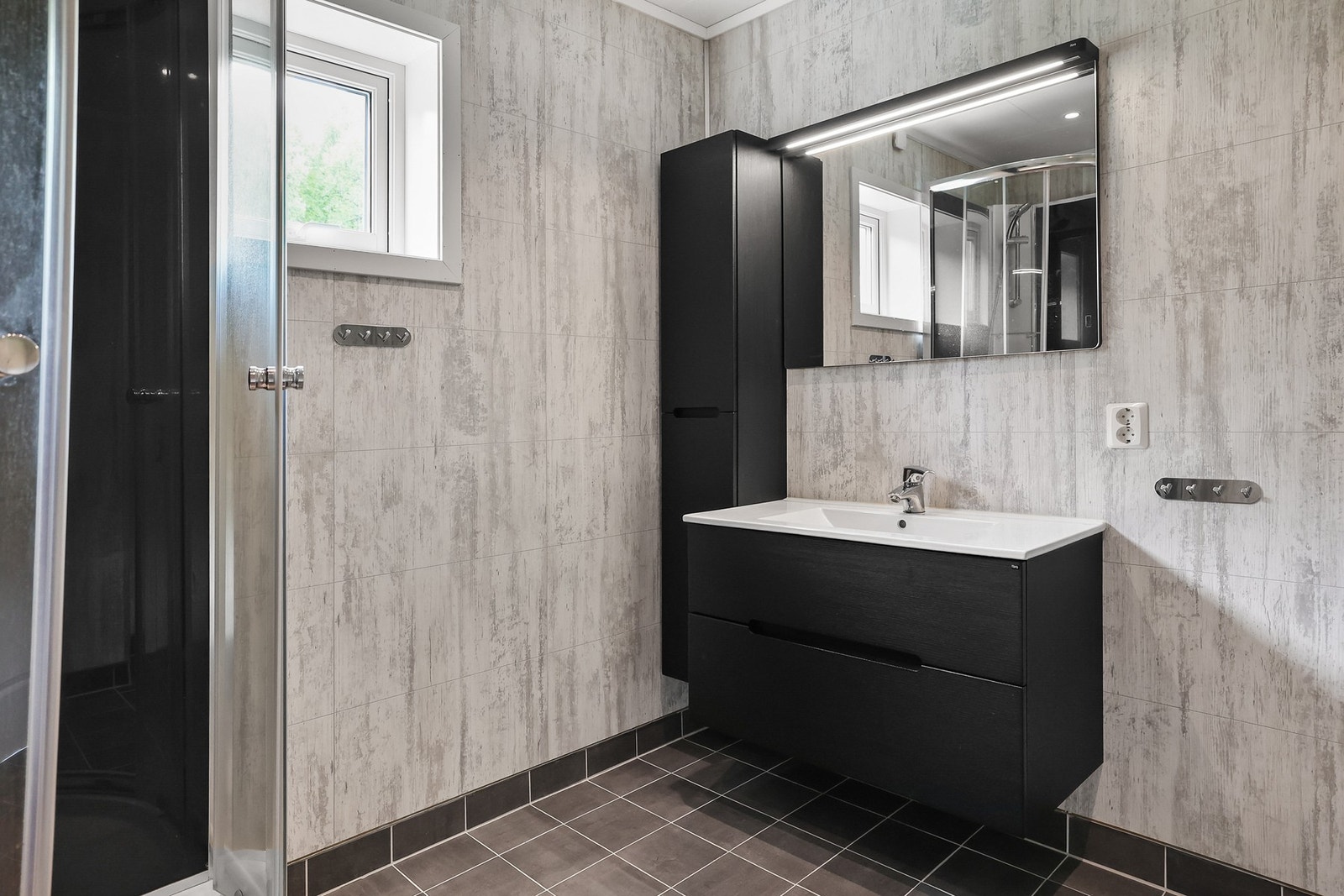 Det er en sort baderomsinnredning med skuffer og nedfelt servant, vegghengt baderomsmøbel med plass til oppbevaring, veggmontert toalett og dusjkabinett. Galleribilde