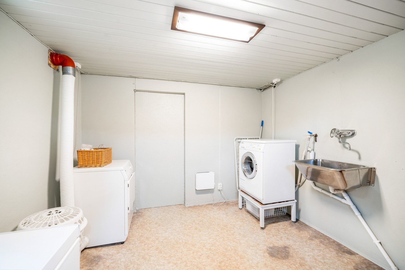 Vaskekjeller med vegghengt utslagsvask i metall og opplegg for vaskemaskin og tørketrommel. Malt panel i himling, belegg på gulvet og malte murvegger. Rommet er ikke bygget eller vurdert som et våtrom da det blant annet ikke er etablert membran/tettesjikt Galleribilde