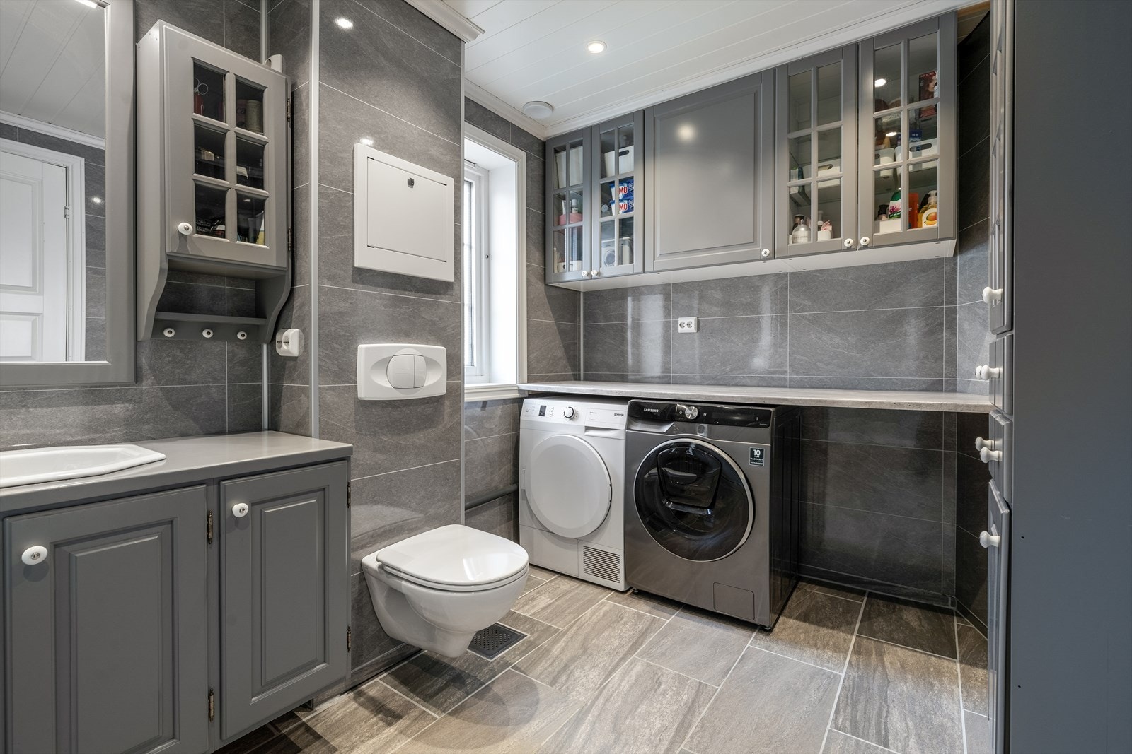 Vaskeromsdelen har en praktisk arbeidsbenk hvor vaskemaskin og tørketrommel er plassert under. Takhøye overskap gir plass til vaskeartikler og gjør dette til et godt arbeidsrom med orden og system. Galleribilde