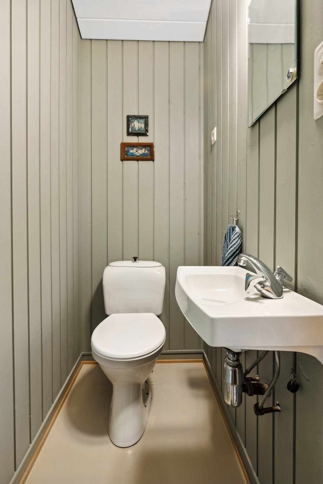 Toalettrom med belegg på gulv, innredet med enkel servant, speil og belysning, samt et toalett. Galleribilde