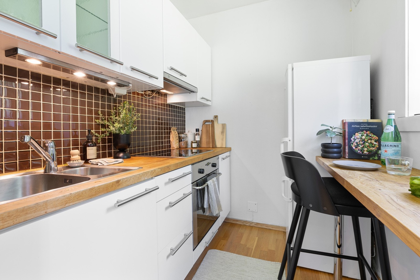 Heltre eik benkeplater med nedfelt oppvaskkum. Lys under overskap ved vask. Fliser på vegg mellom overskap og kjøkkenbenk. Galleribilde