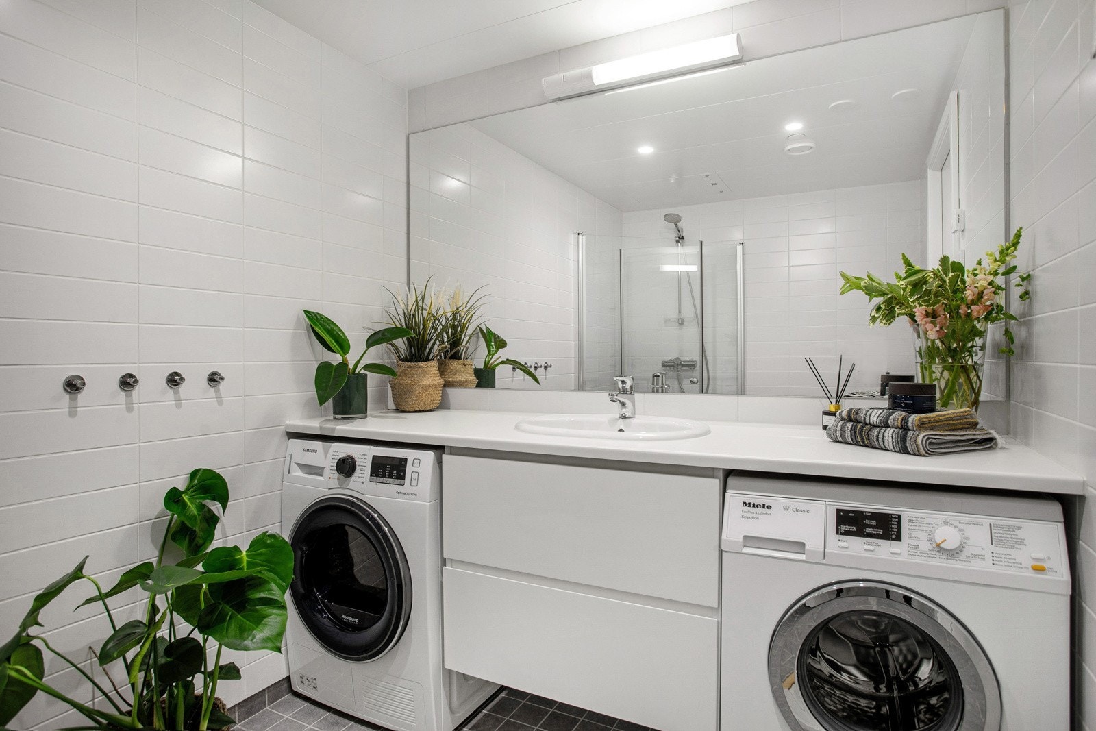 Moderne baderomsinnredning med skuffer og laminert benkeplate med nedsenket servant, stort speil med belysning over. Praktisk med plass og både vaskemaskin og tørketrommel under benkeplaten. Galleribilde