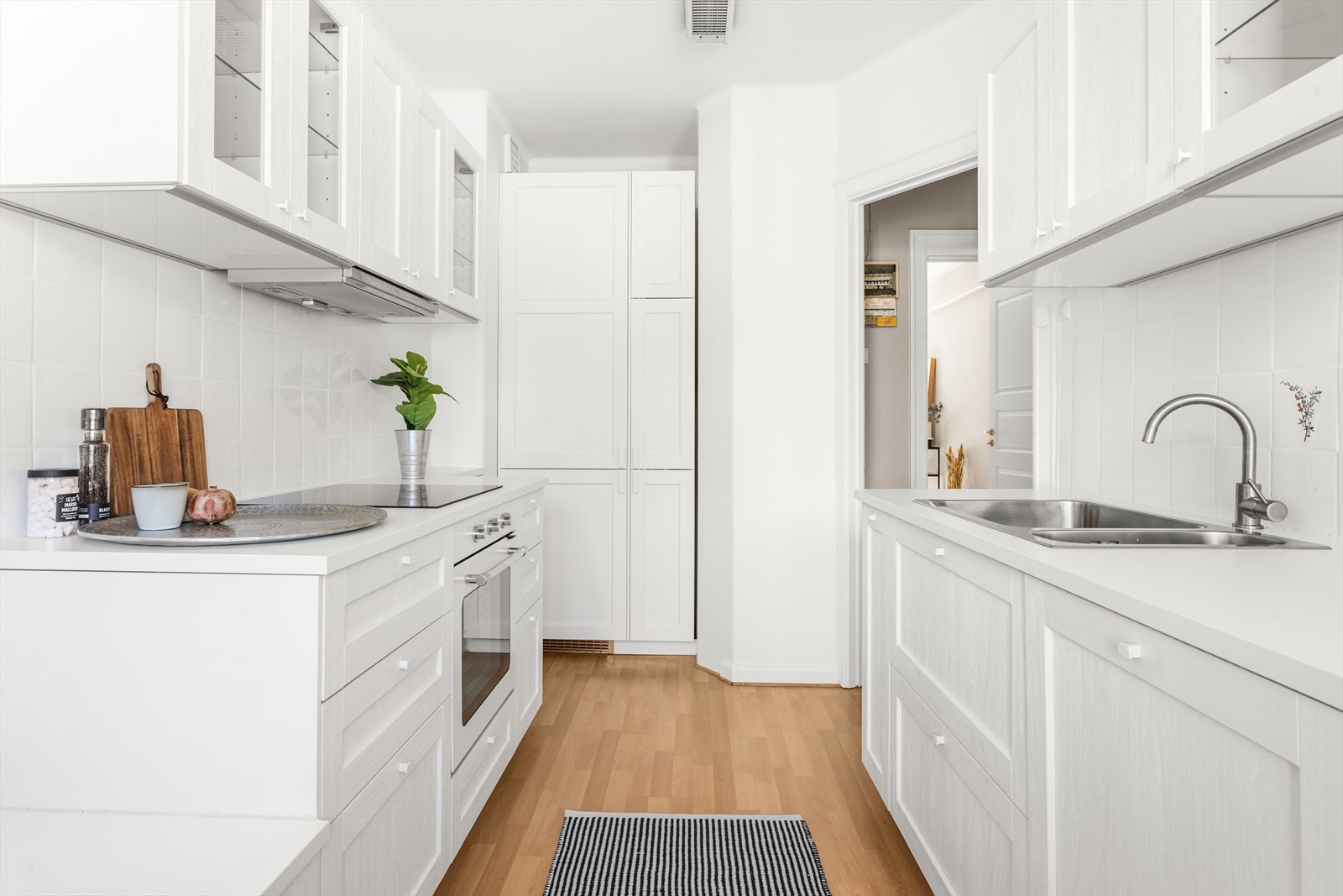 Kjøkkeninnredning med hvite profilerte, folierte fronter samt noen vitrinedører, laminat benkeplate med nedfelt kum i rustfritt stål og ettgreps blandebatteri, lys under overskap og fliser på vegg over benkeplaten. Galleribilde