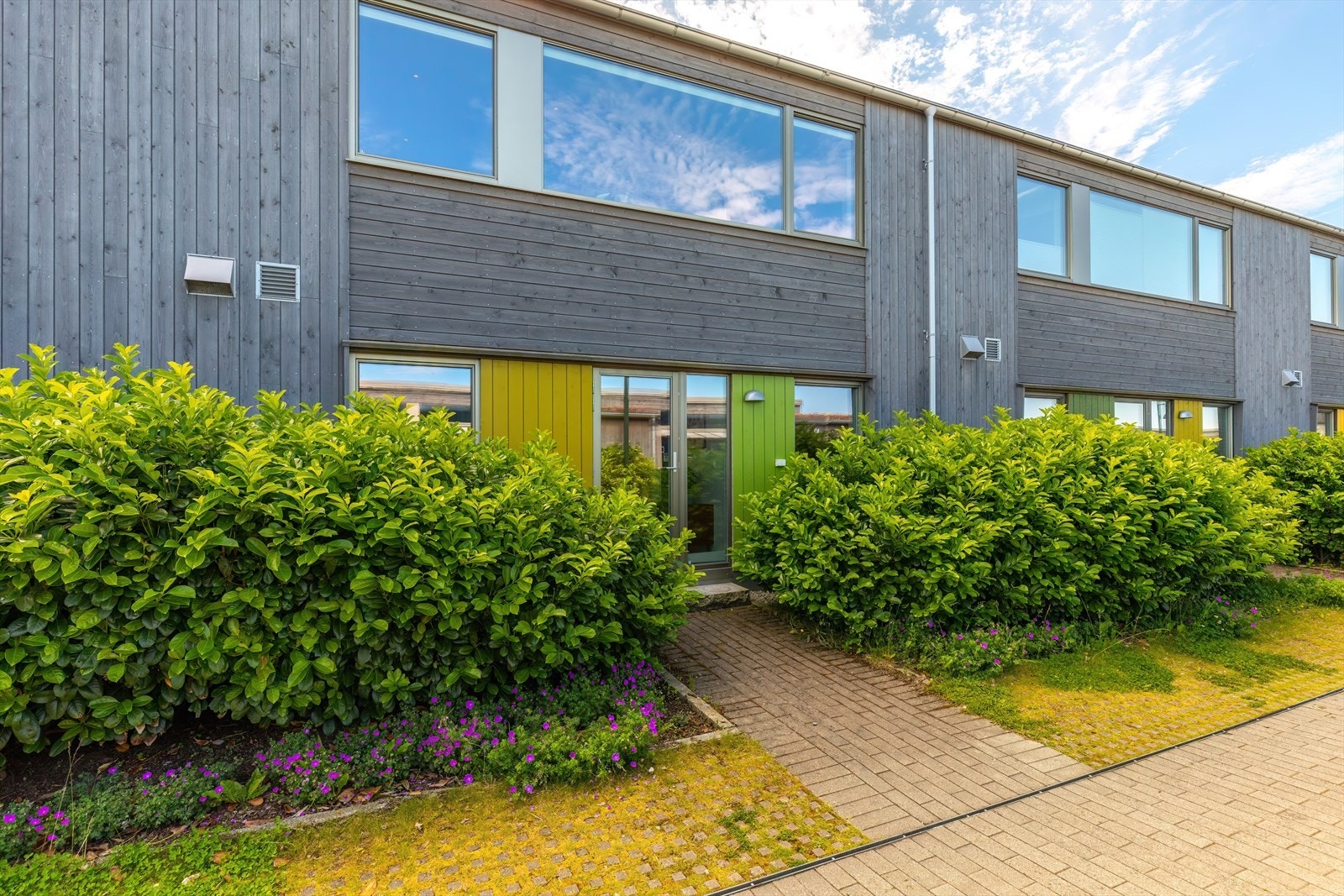 Utvendig inngangsparti fra nedre siden er belagt med belegningsstein og beplantet med busker og blomster. Galleribilde