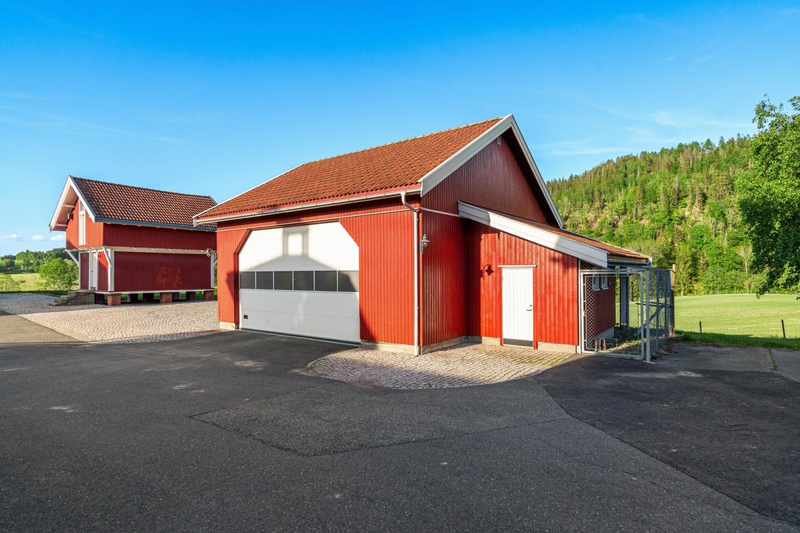 Asfaltert- og brostensbelagt gårdsplass med dreneringskum. Her med stor dobbelt garasje m/automatisk port, hønsehus m/utegård samt et gammelt stabbur som ble renovert i 2007. Galleribilde