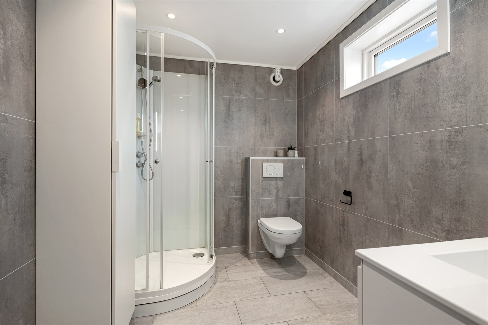 Bad i kjeller med flislagt gulv med varmekabler, baderomsplater på vegger. Servantinnredning, veggskap, dusjkabinett og vegghengt wc. Galleribilde