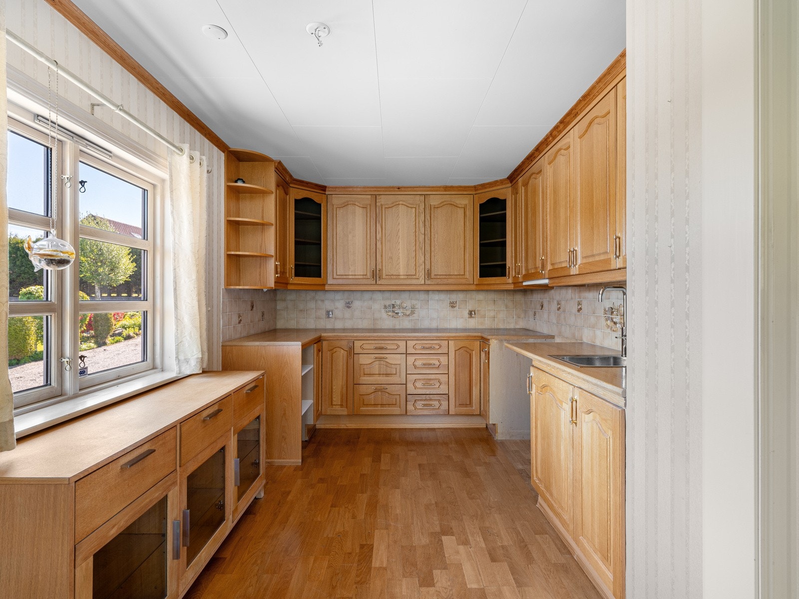 Kjøkkenet ligger i en åpen løsning med stuen, men er likevel skjermet fra deler av rommet. Kjøkkeninnredningen består av gylne, profilerte fronter i heltre samt benkeplate i tre. Galleribilde