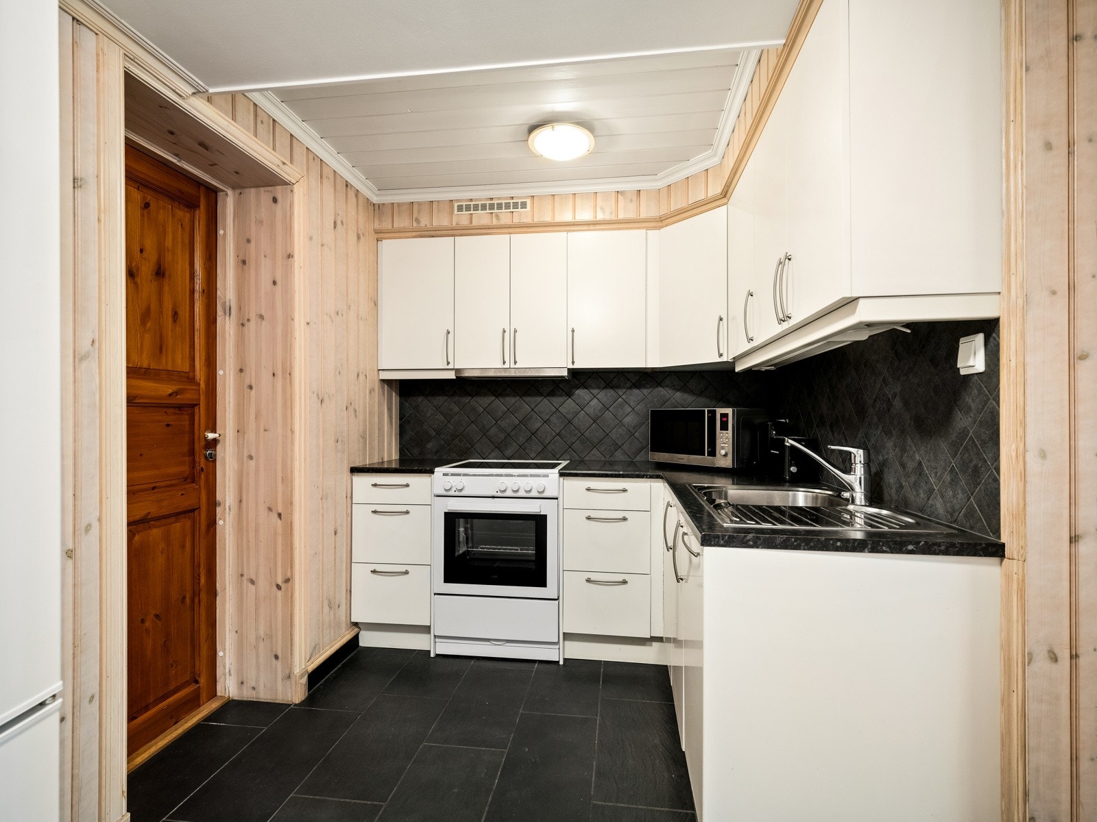 Kjøkkeninnredningen har lyse, glatte fronter og laminert benkeplate med nedfelt oppvaskkum. Det er montert mørke fliser på veggen mellom benk og overskap. Det er lys under overskapene, og over kokesonen er det ventilator. Galleribilde