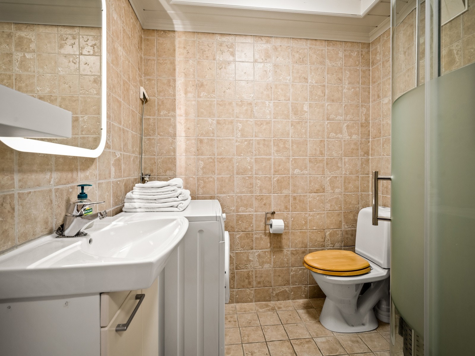 Badet har fliser på gulvet, fliser på veggene, gulvvarme, gulvmontert toalett, dusjhjørne og opplegg for vaskemaskin. Innredningen består av en heldekkende servant med underskap, og over servanten er det speil med integrert belysning. Galleribilde