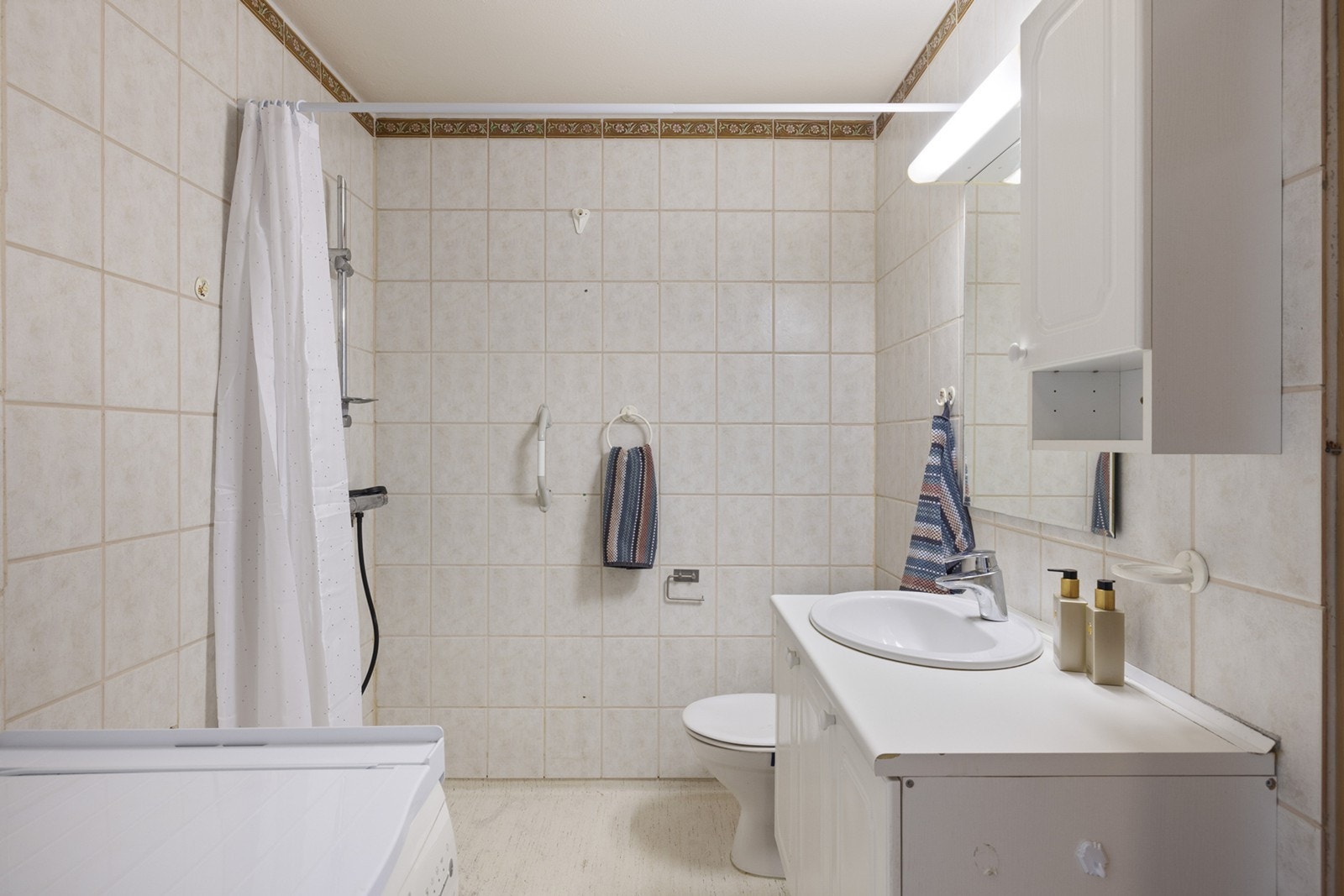 Stort bad med gulvbelegg på gulv og fliser på vegger. Innredningen består av et underskap/overskap, servant, speil og armatur med stikk. Badet vil bli totalrenovert av borettslaget i nær fremtid. Se salgsoppgaven for mer informasjon. Galleribilde