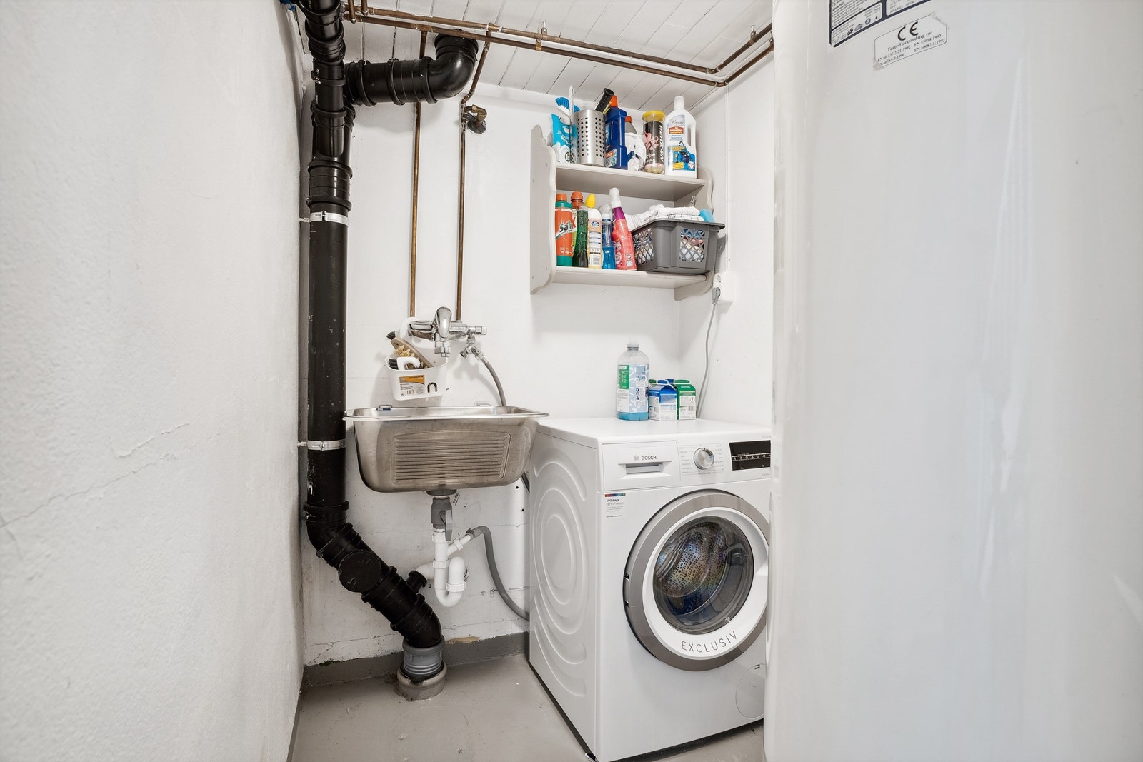 Vaskerommet har hvitmalt betong på vegger og gråmalt betong på gulvet. Rommet er innredet med utslagsvask på vegg og opplegg for vaskemaskin. Naturlig ventilering fra rommet. Galleribilde