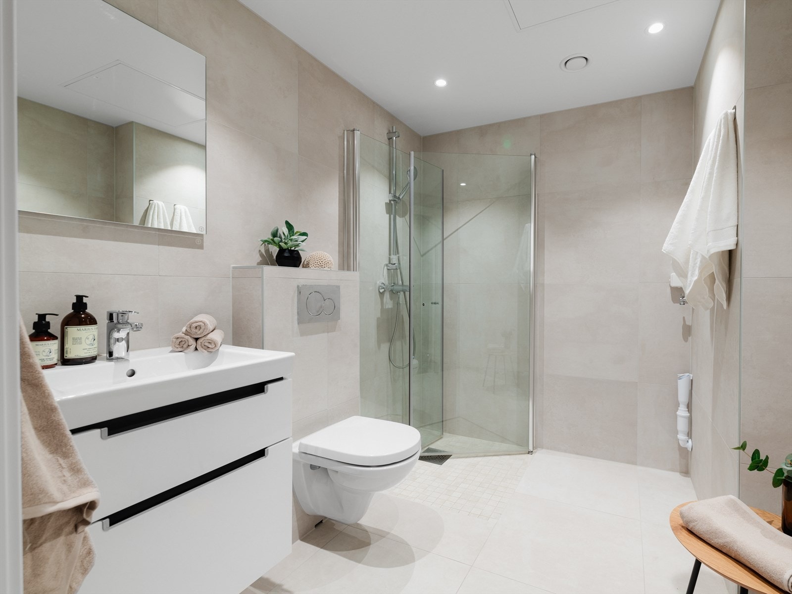 Badet er av god størrelse. Rommet er delikat flislagt med lyse sandgrå fliser, og innredet med dusjhjørne med innfellbare glassdører, vegghengt klosett, servantinnredning og speil. Galleribilde