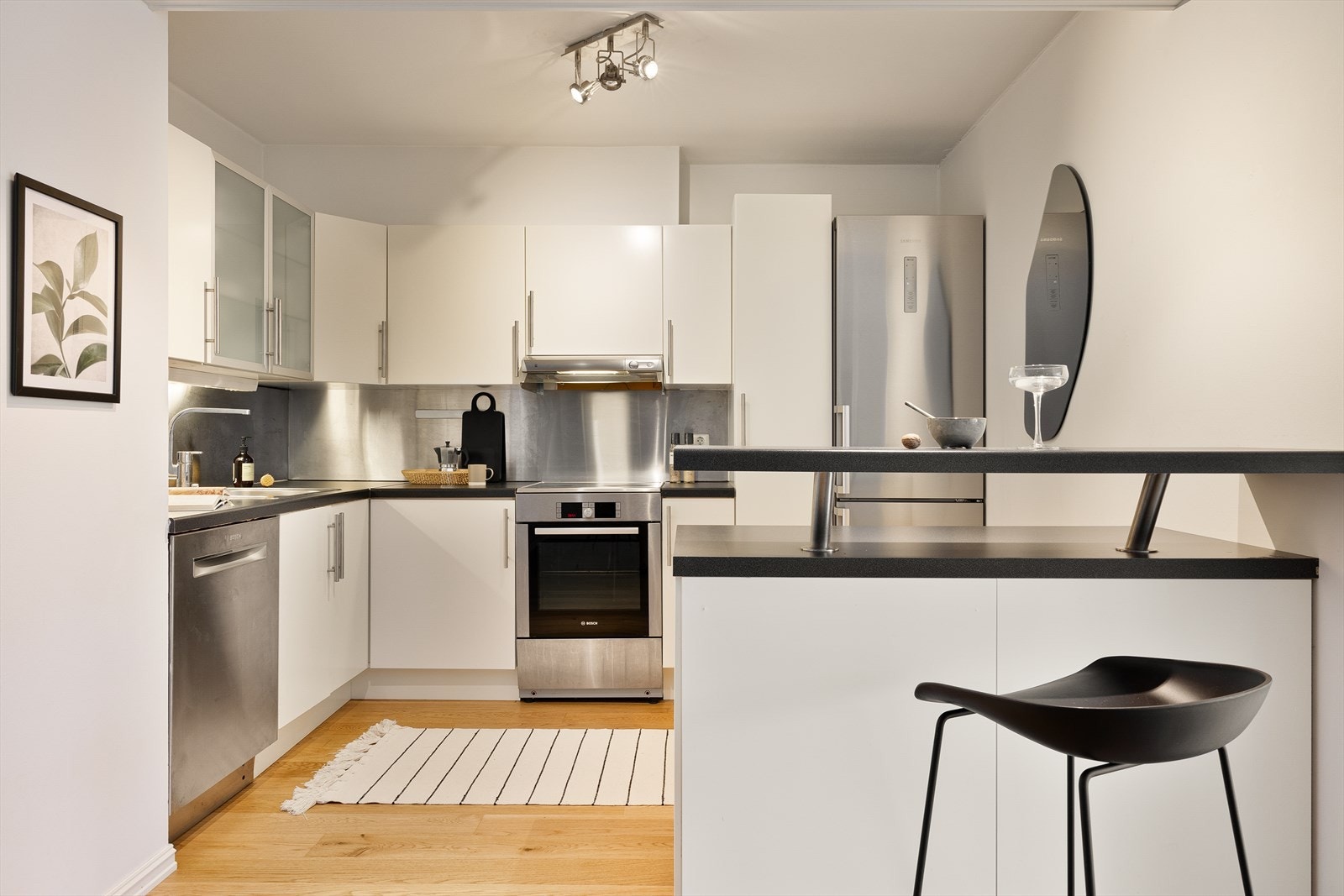 Lys og tidløs kjøkkeninnredning med slette fronter og laminat benkeplate. Ett av overskapene har glassfront, over benkeplaten er det benyttet plate, det er stål oppvaskkum og ventilator. Galleribilde