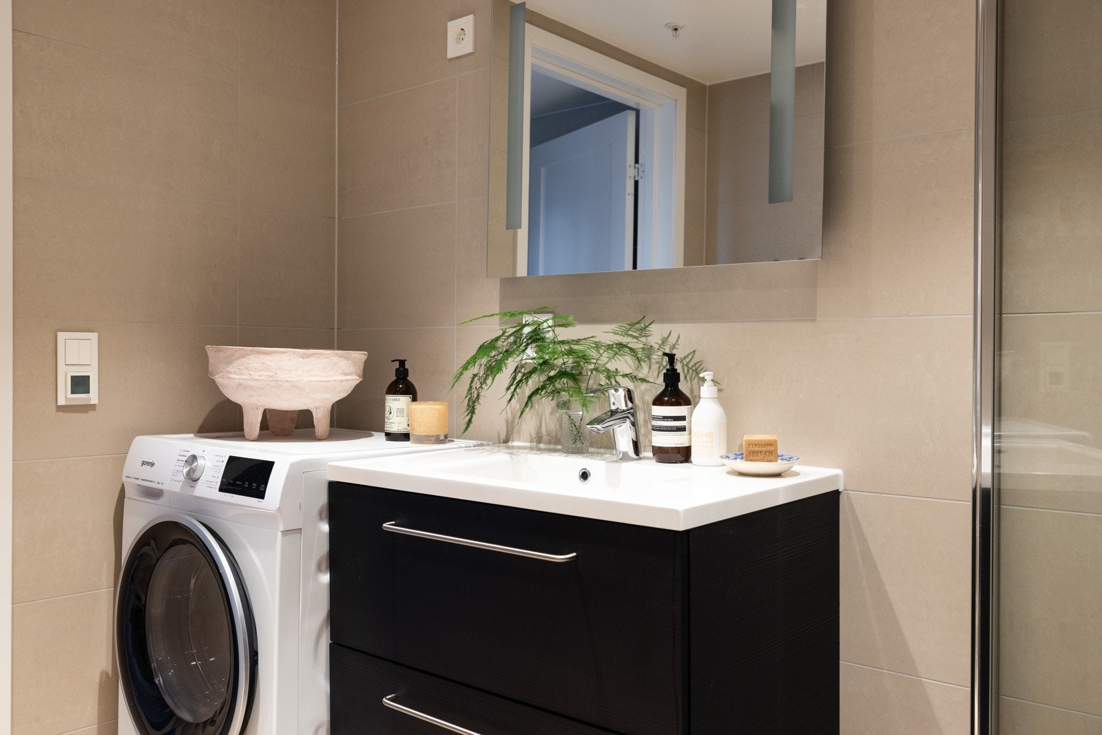 Badet er innredet med dusjnisje med glassdører, nedsenket håndvask med skuffer og speil, veggmontert klosett og opplegg til vaskemaskin. Galleribilde