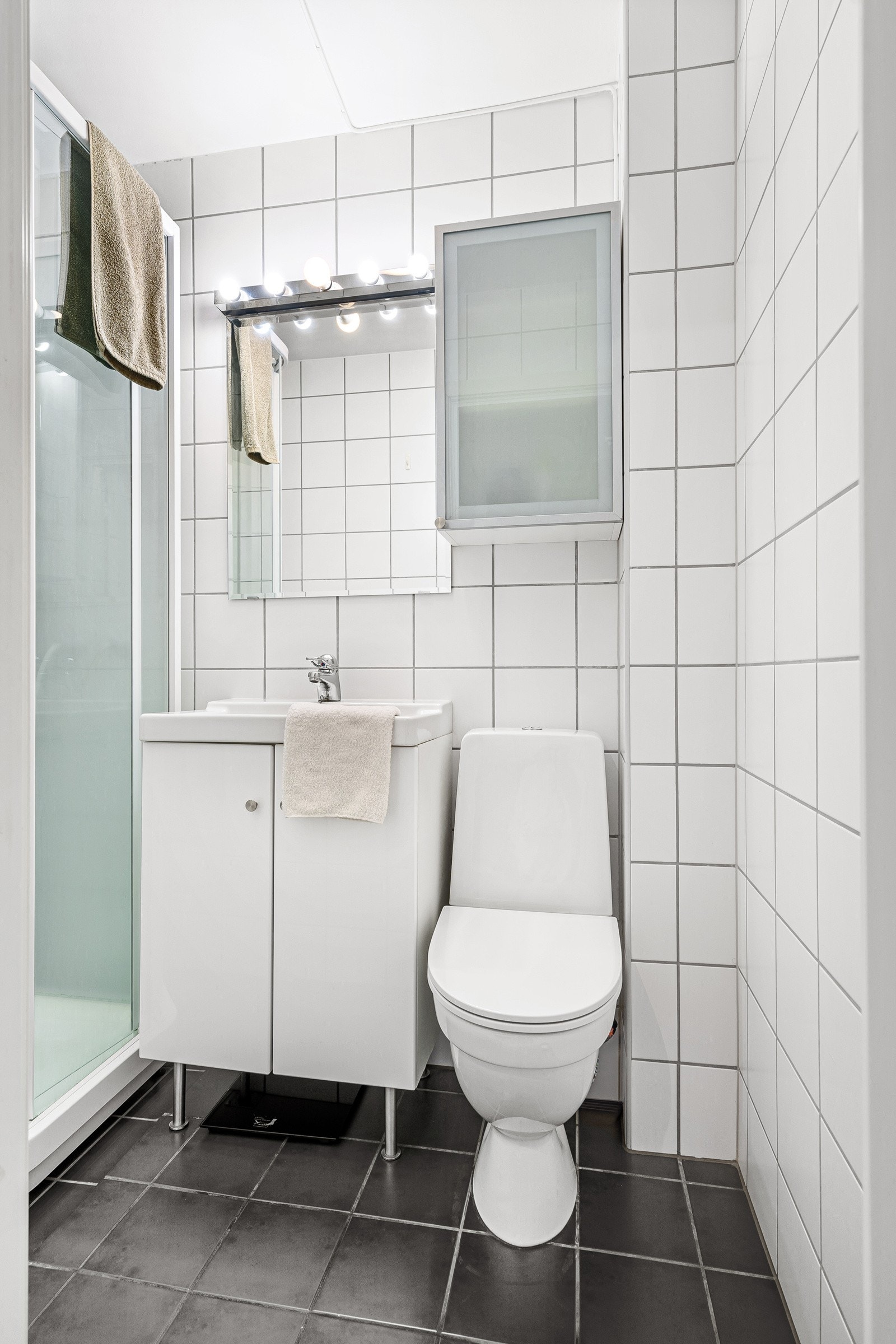 Badet er innredet med dusjkabinett, vegghengt belysning over speil og servant, og gulvstående toalett. Galleribilde