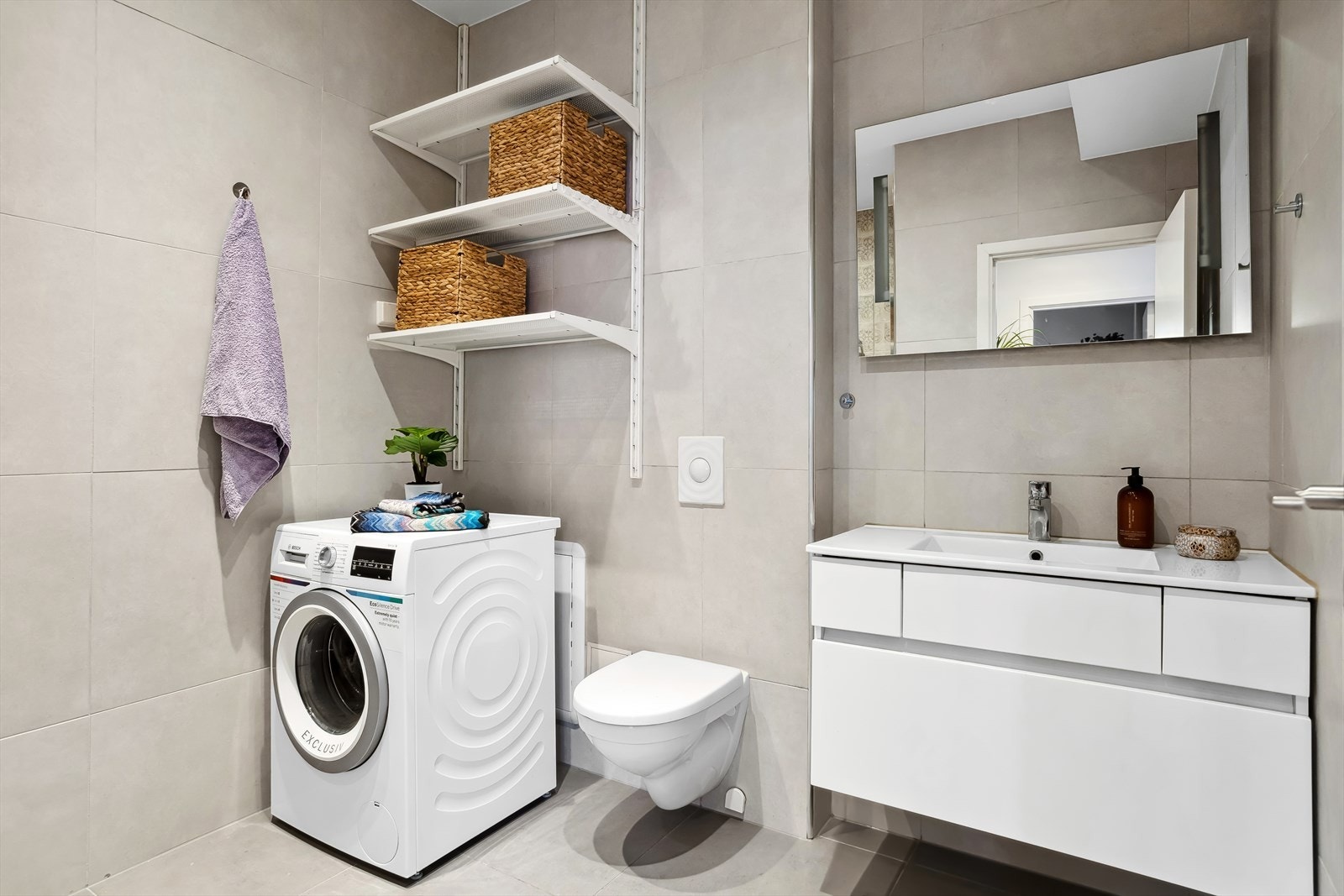 Badet er romslig og delikat med grå, pene fliser med varme i gulv. Det er downlights i himling og ekstra oppbevaringsplass til tøymykner, vaskepulver og håndklær på vegghengte hyller. Galleribilde