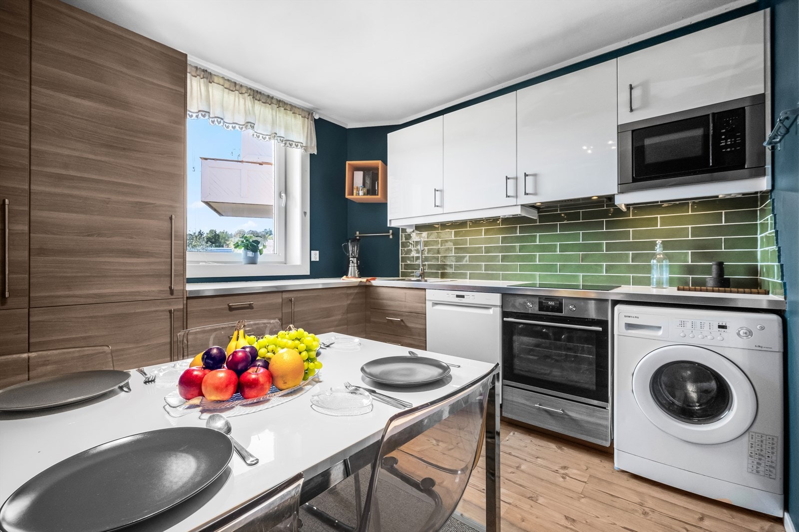 Belysning under overskap. Fliser på vegg over benkeskap. Laminat benkeplate med nedfelt kum i rustfritt stål. Kullfilterventilator. Induksjonstopp, stekeovn og kjøl/frys er integrert i innredningen. Opplegg for oppvaskmaskin og vaskemaskin. Galleribilde