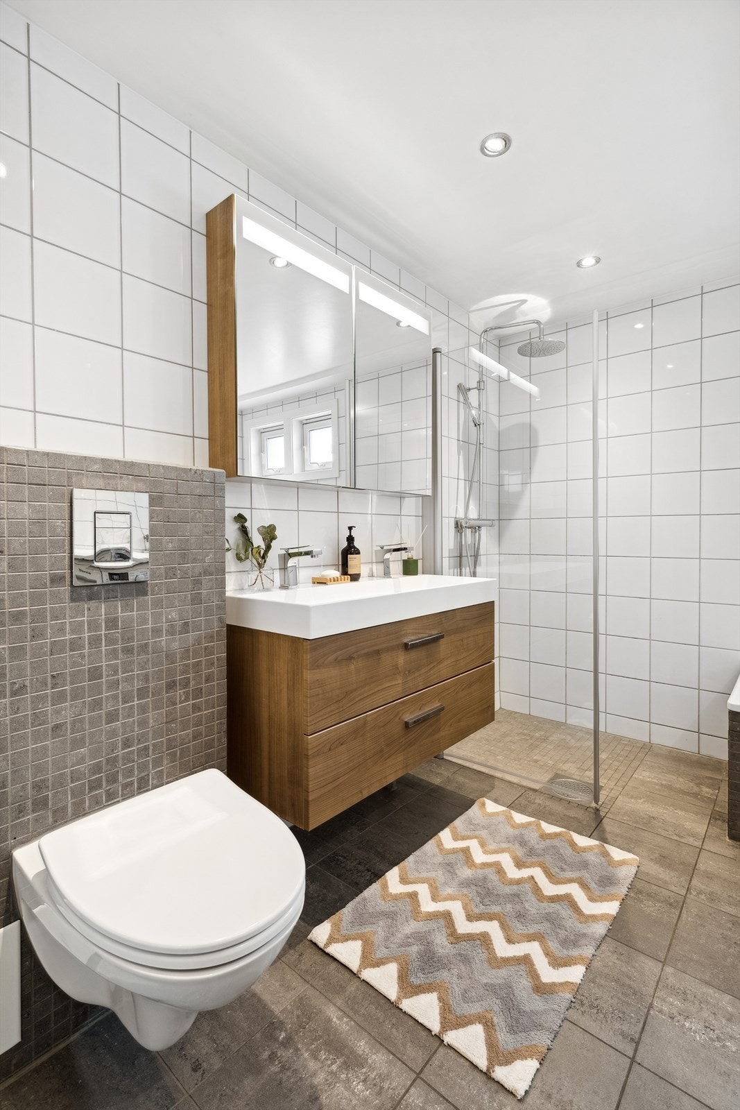 Innredning bestående av servantskap med glatte fronter, overskap med speildører med integrert LED-lys, høyskap, vegghengt wc, innebygd badekar med flislagt front og dusjhjørne med svingdør. Galleribilde