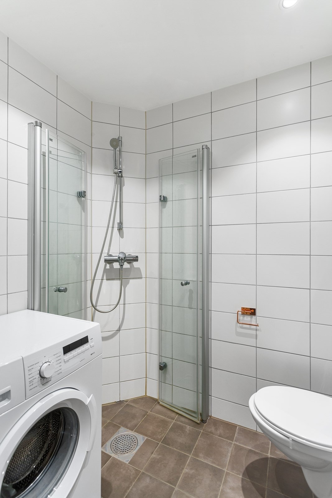 Badet er utstyrt med dusjsone med innfellbare dører, veggmontert wc, servantskap med ett-greps blandebatteri og opplegg for vaskemaskin Galleribilde
