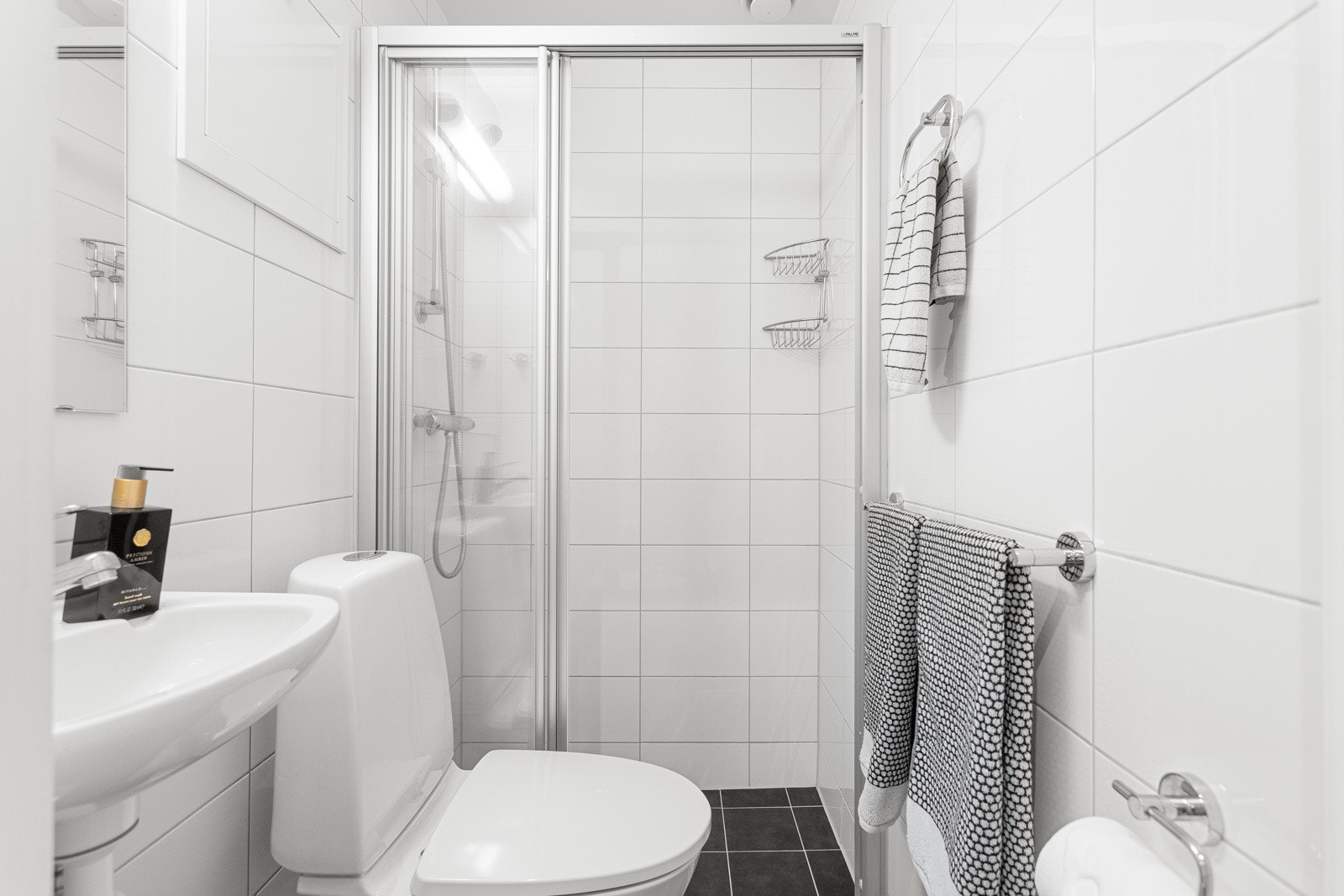 Baderommet har mørke fliser på gulv med gulvvarme, hvite fliser på veggene og mekanisk avtrekksventil i himling. Galleribilde