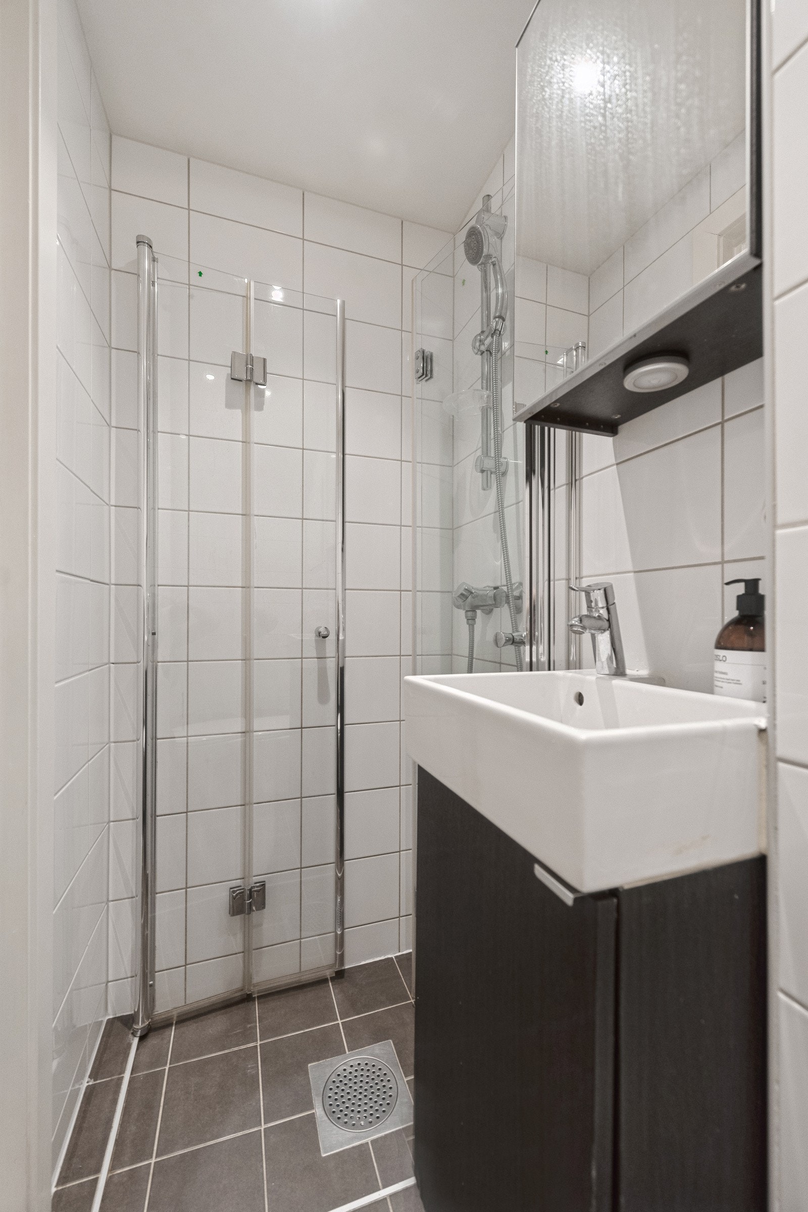 Badet inneholder gulvstående servantinnredning med dør og ovenpåliggende servant med ett-greps armatur, speilskap med lys og stikkontakt på vegg over servant, dusjhjørne med glassdører, veggmontert dusjarmatur og vegghengt toalett. Galleribilde