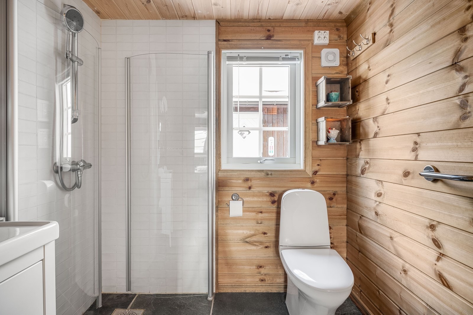 Badet er utstyrt med veggmontert dusjbatteri og innfellbare dusjvegger, gulvmontert toalett og baderomsinnredning med skuffer og heldekkende servant. Galleribilde