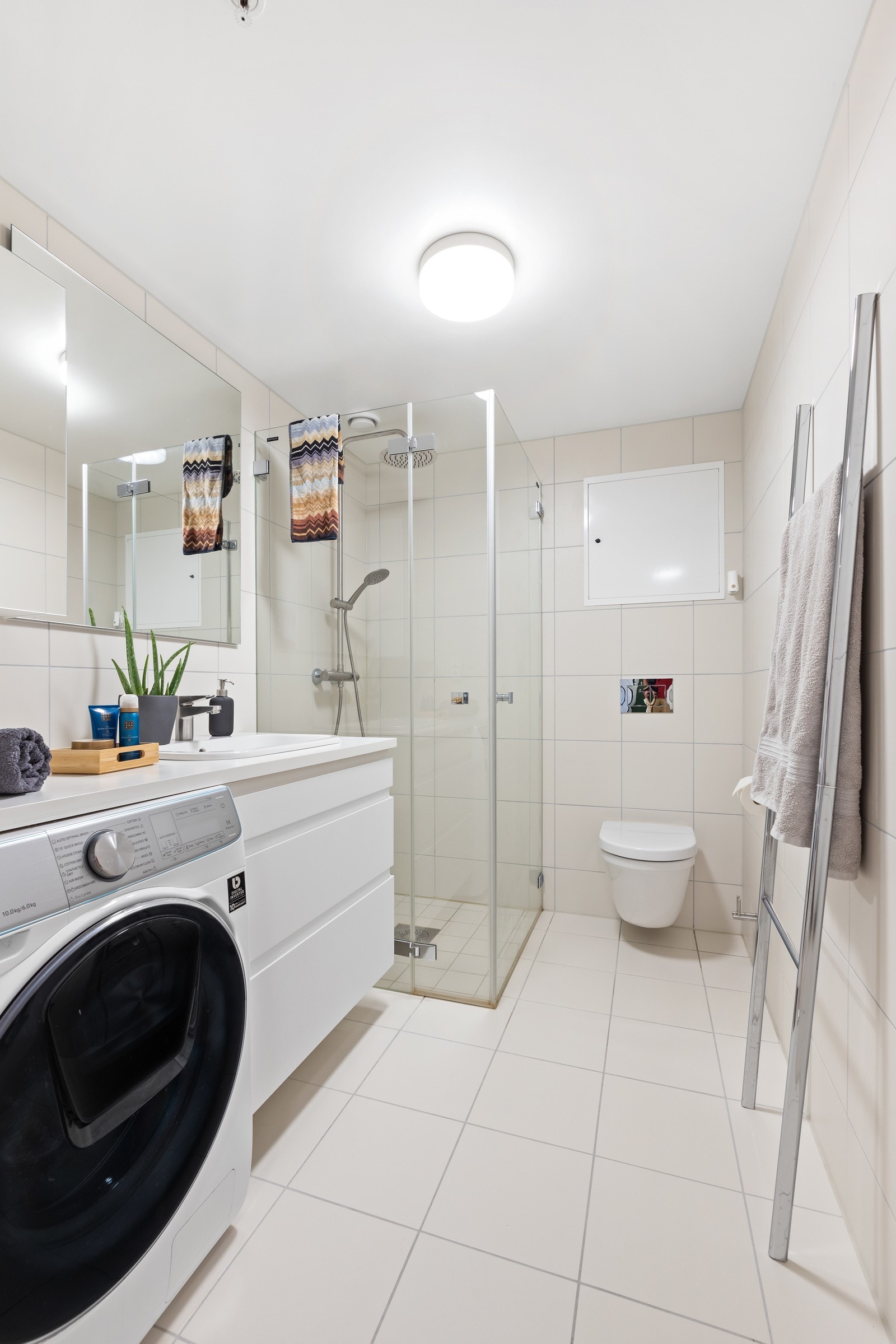 Flislagt baderom innredet med dusjnisje, servant med skuffeseksjon, vegghengt klosett og opplegg for vaskemaskin. Galleribilde