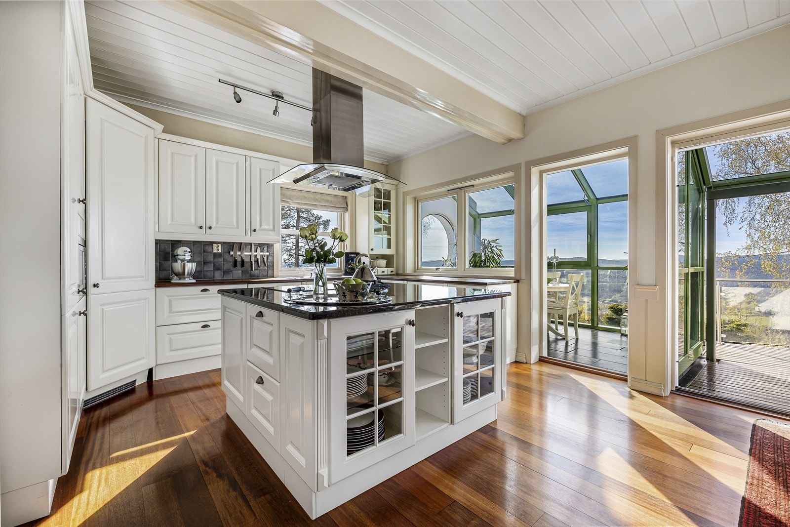 Kjøkken med innredning fra JKE Design. Hvite profilerte fronter på skuffer og skap og heltre benkeplate. Stor kjøkkenøy i samme utførelse og kvalitet som resten av innredningen, men med steinplate Galleribilde