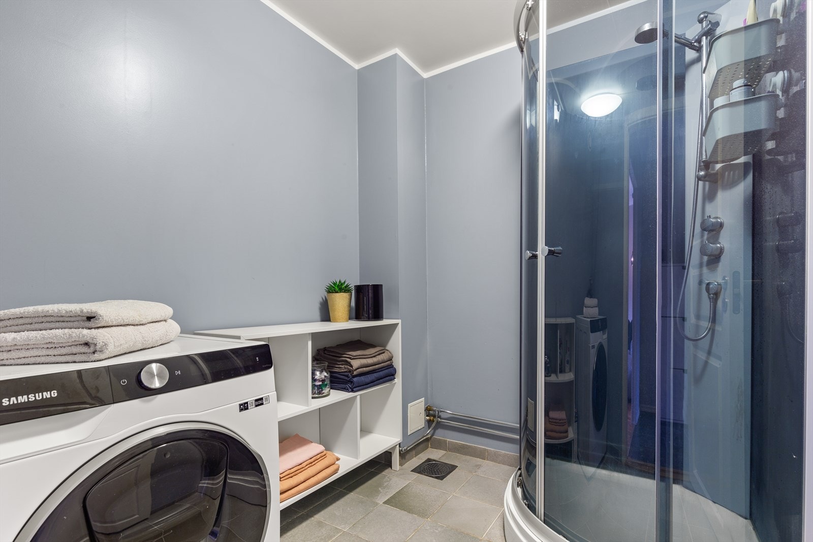 Vaskerommet har grå fliser på gulv, sluk, malte vegger, dusjkabinett samt opplegg for vaskemaskin. Galleribilde