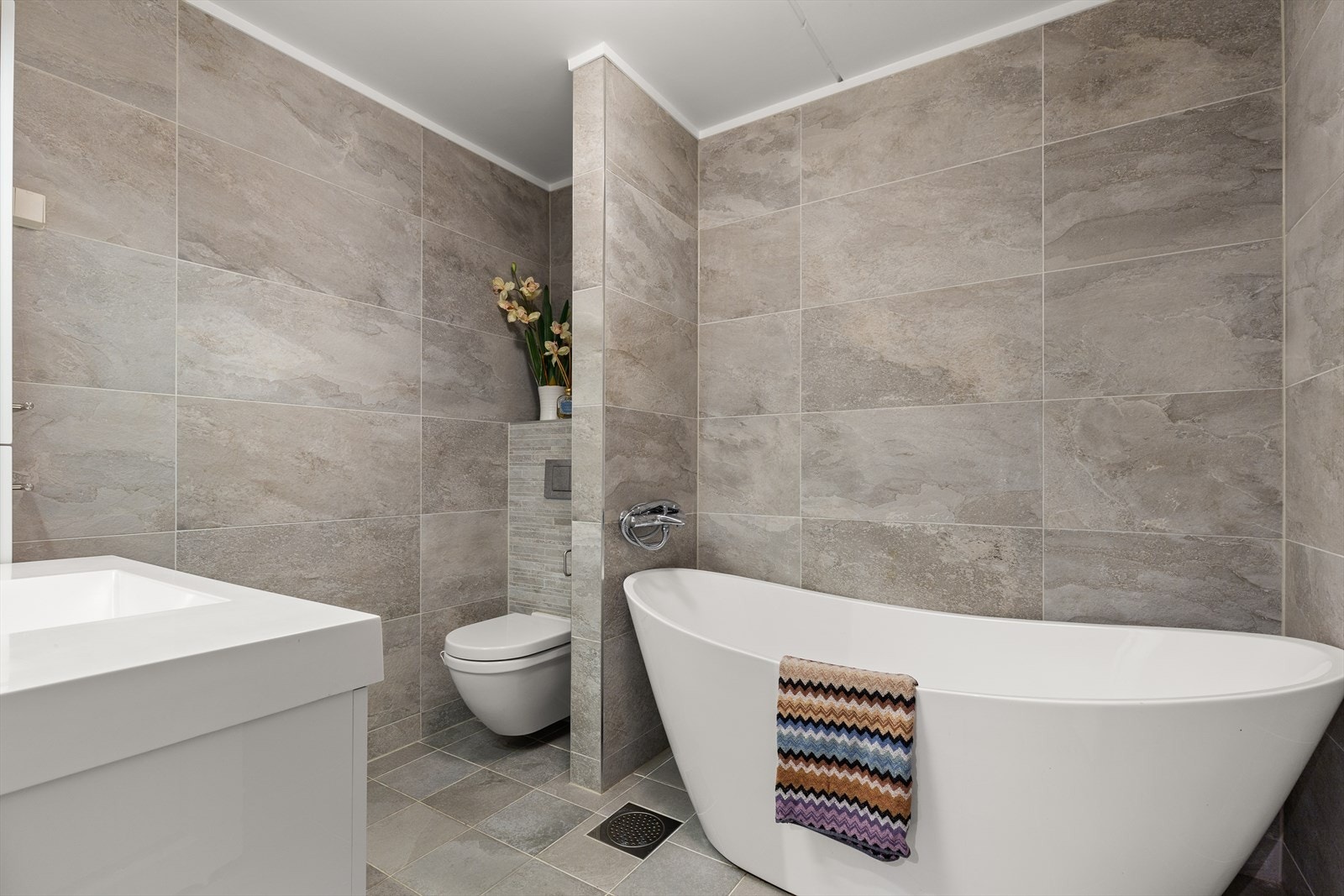 Badet er fra 2016 og har grå fliser på gulv og vegg samt varme i gulv. Innredningen består av servant på hvitt servantskap, matchende sideskap, speil og et frittstående badekar. Galleribilde